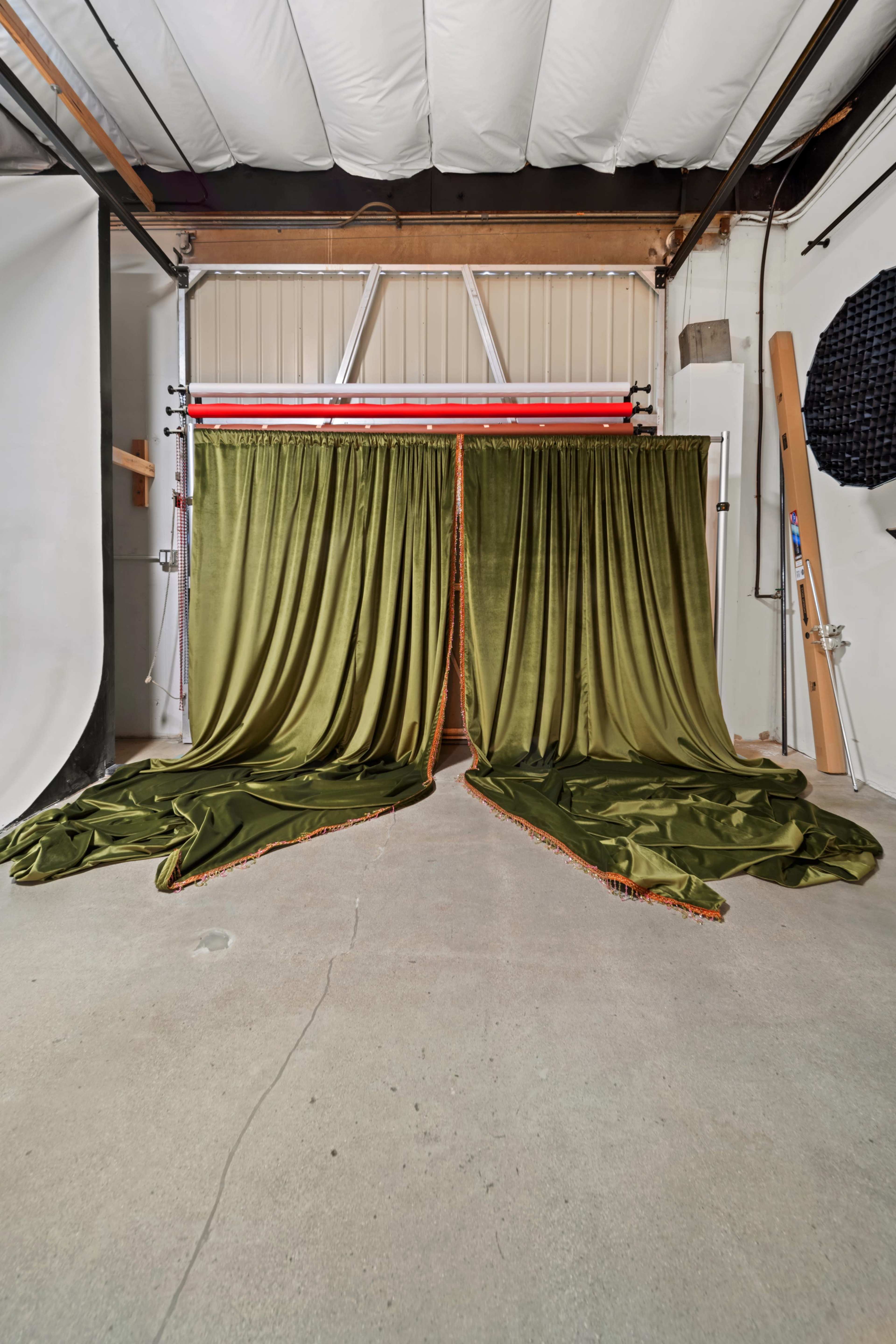 The image shows a studio space with two large, green fabric curtains parted in the center, revealing a concrete floor and various studio equipment in the background.