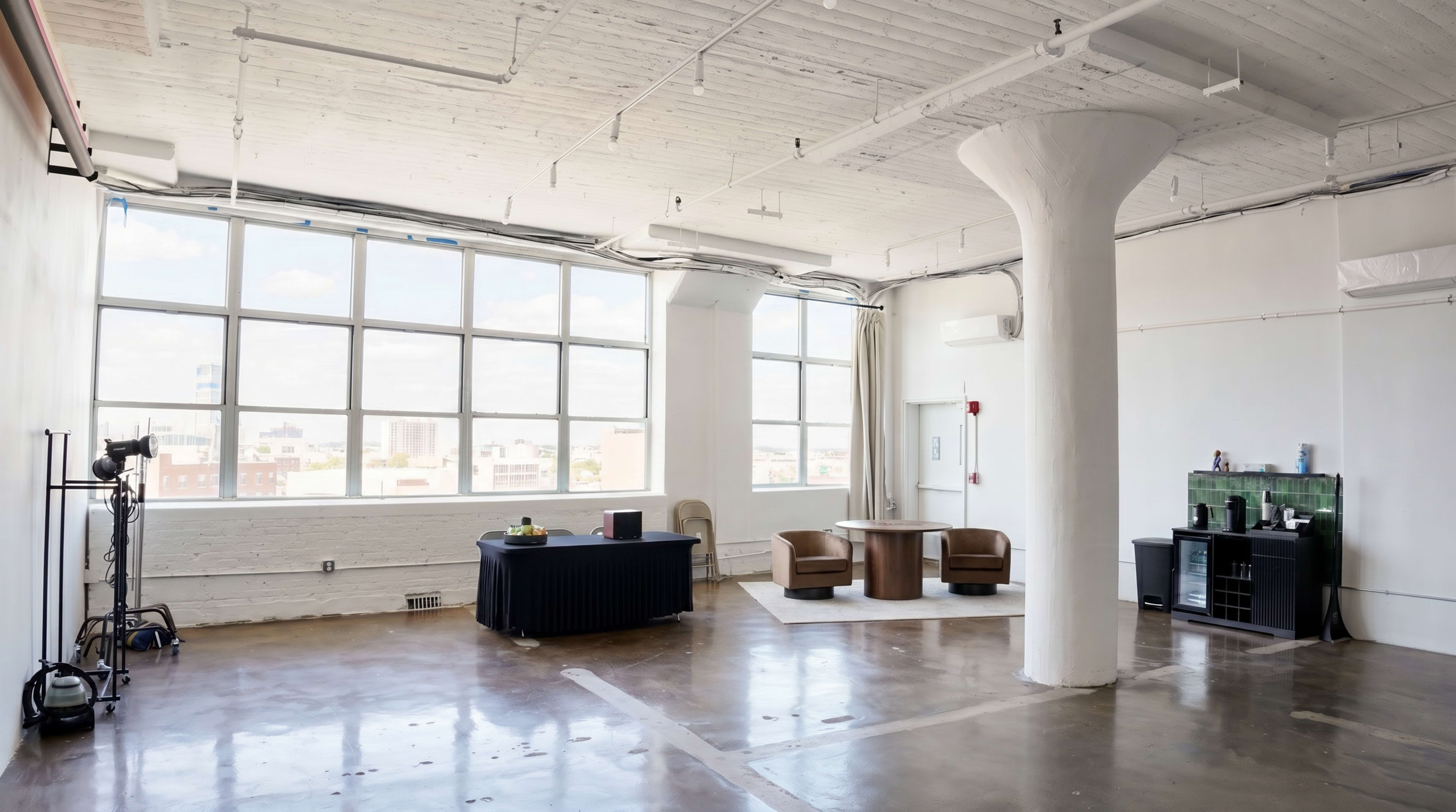 The image depicts a spacious, modern interior with large windows, a table and chairs in the center, and various furnishings along the walls.