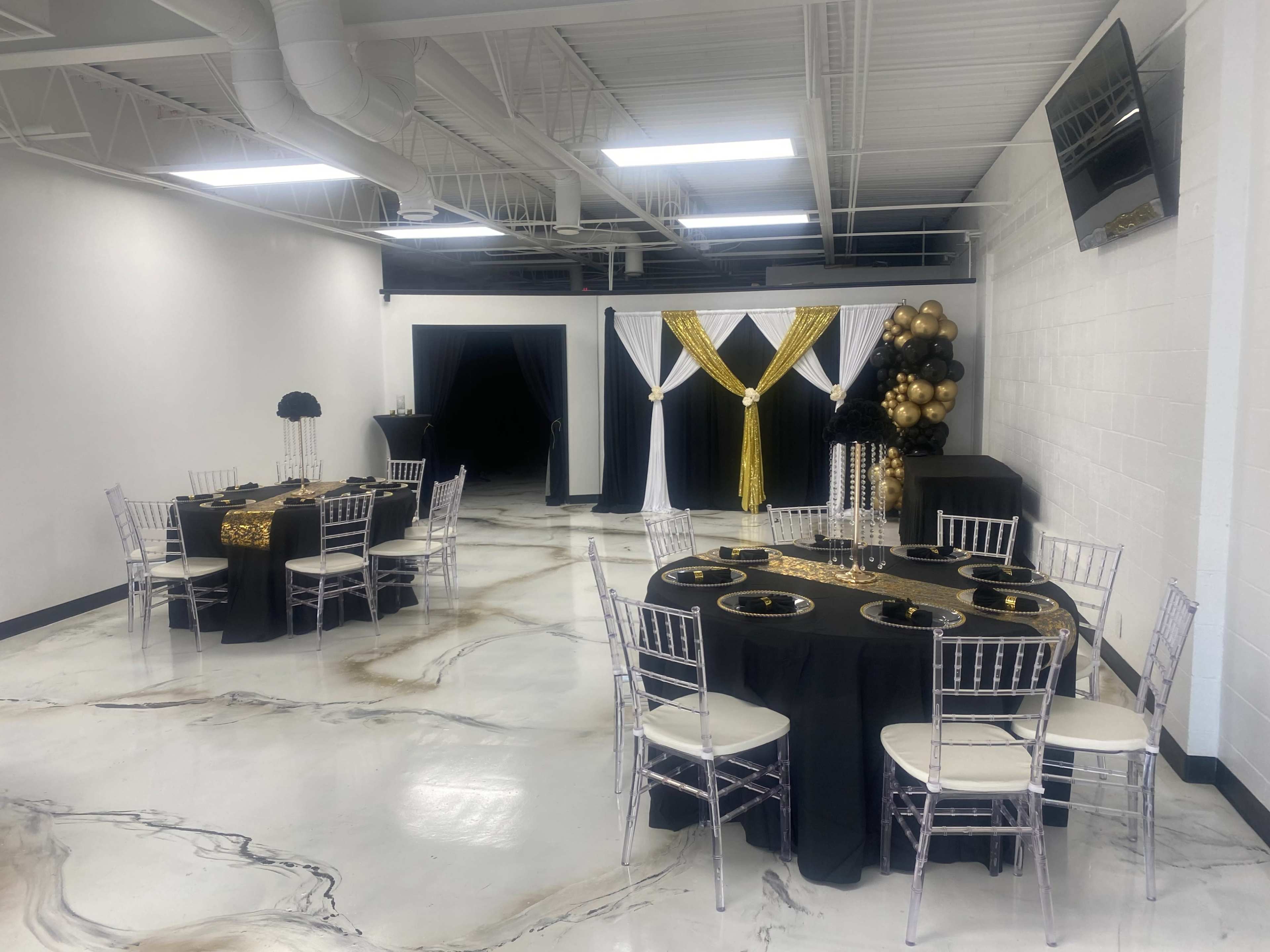 A banquet room with black and gold themed tables set up for an event, featuring elegant table settings and decorative elements like balloons and drapes.