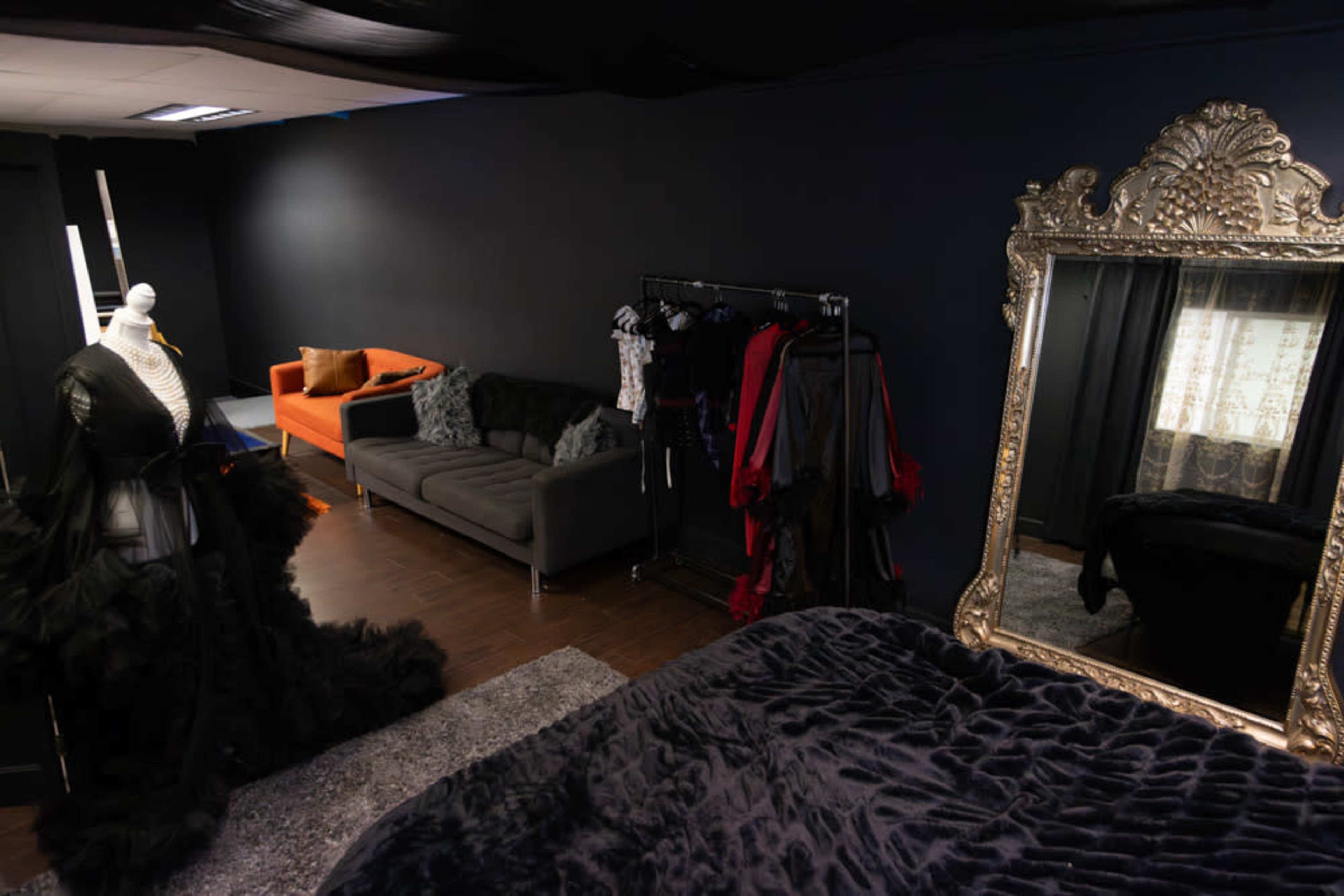A dimly lit studio with a mannequin dressed in a black gown, an orange couch, a clothing rack with various garments, and a large decorative mirror.