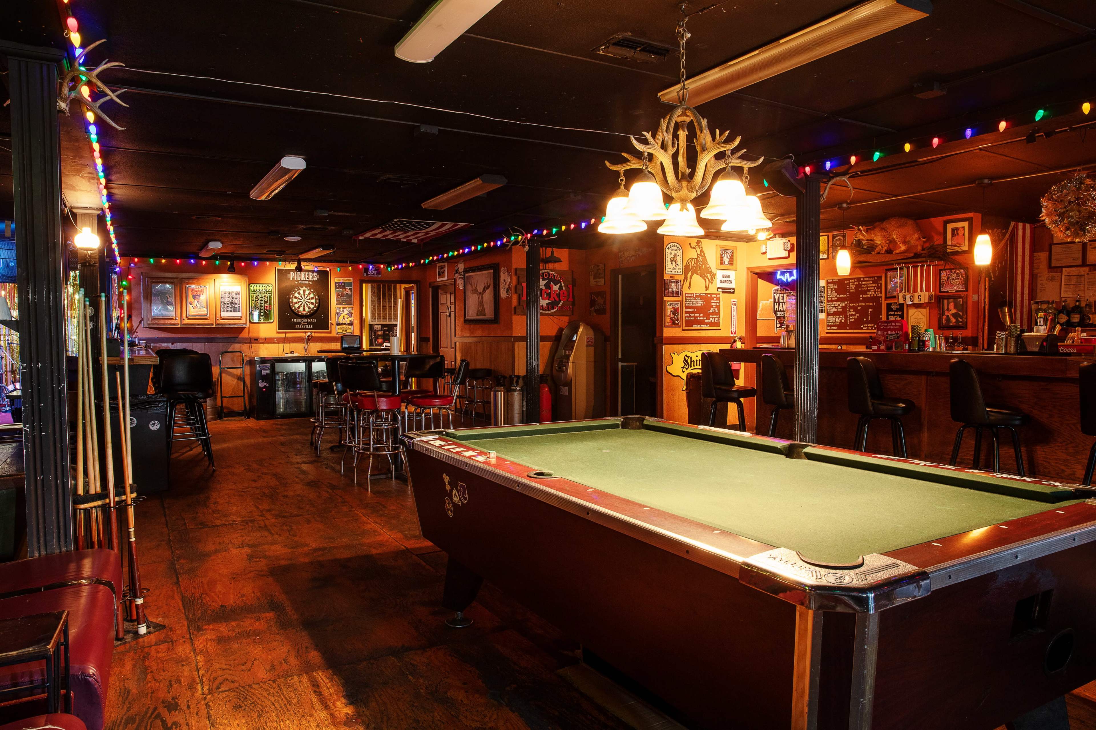 The image shows a dimly lit bar featuring a pool table, colorful string lights, and a wooden counter with stools.