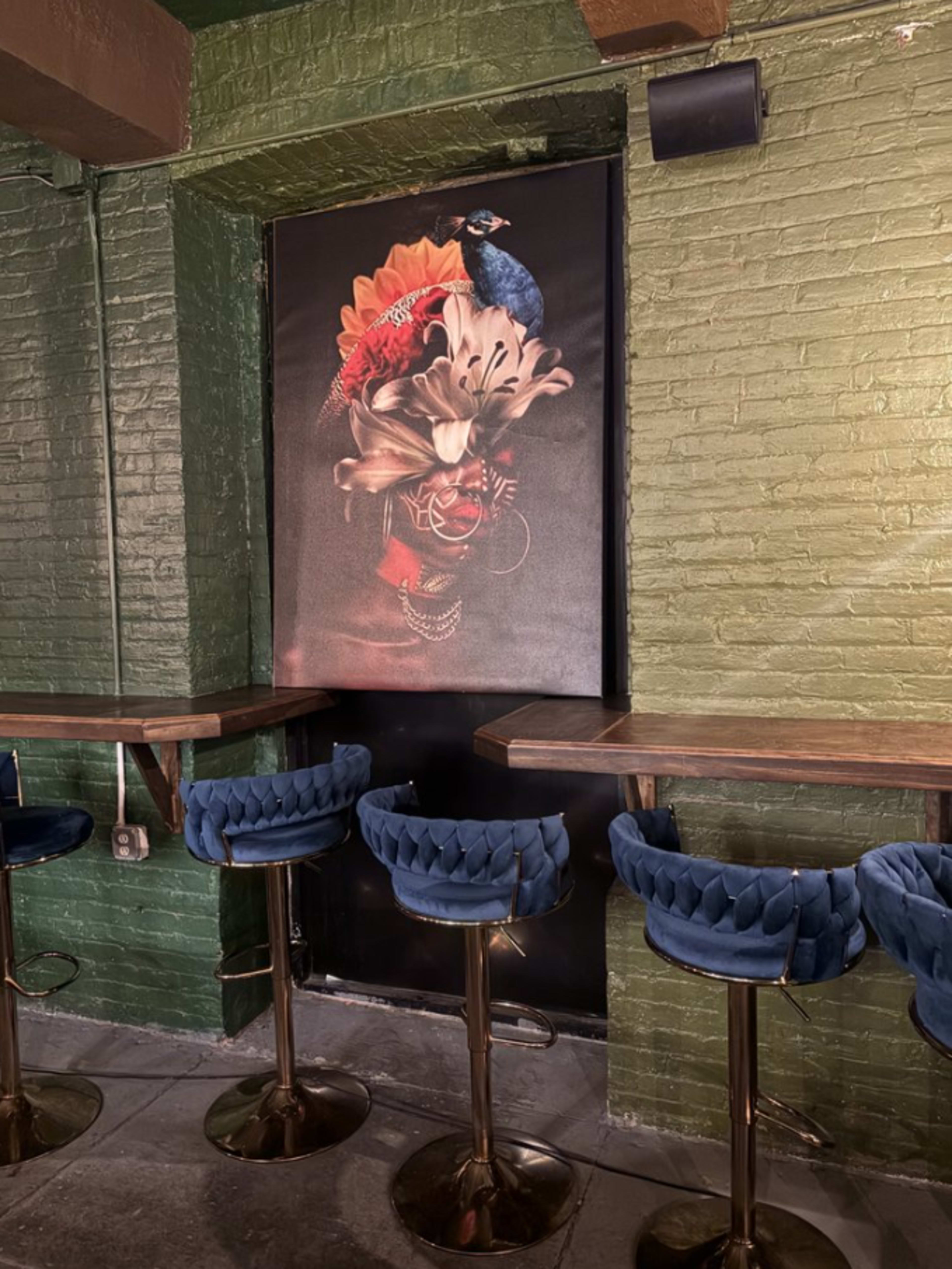 The image shows a stylized mural of a peacock and flowers on a door, surrounded by green brick walls and blue upholstered bar stools along a wooden countertop.