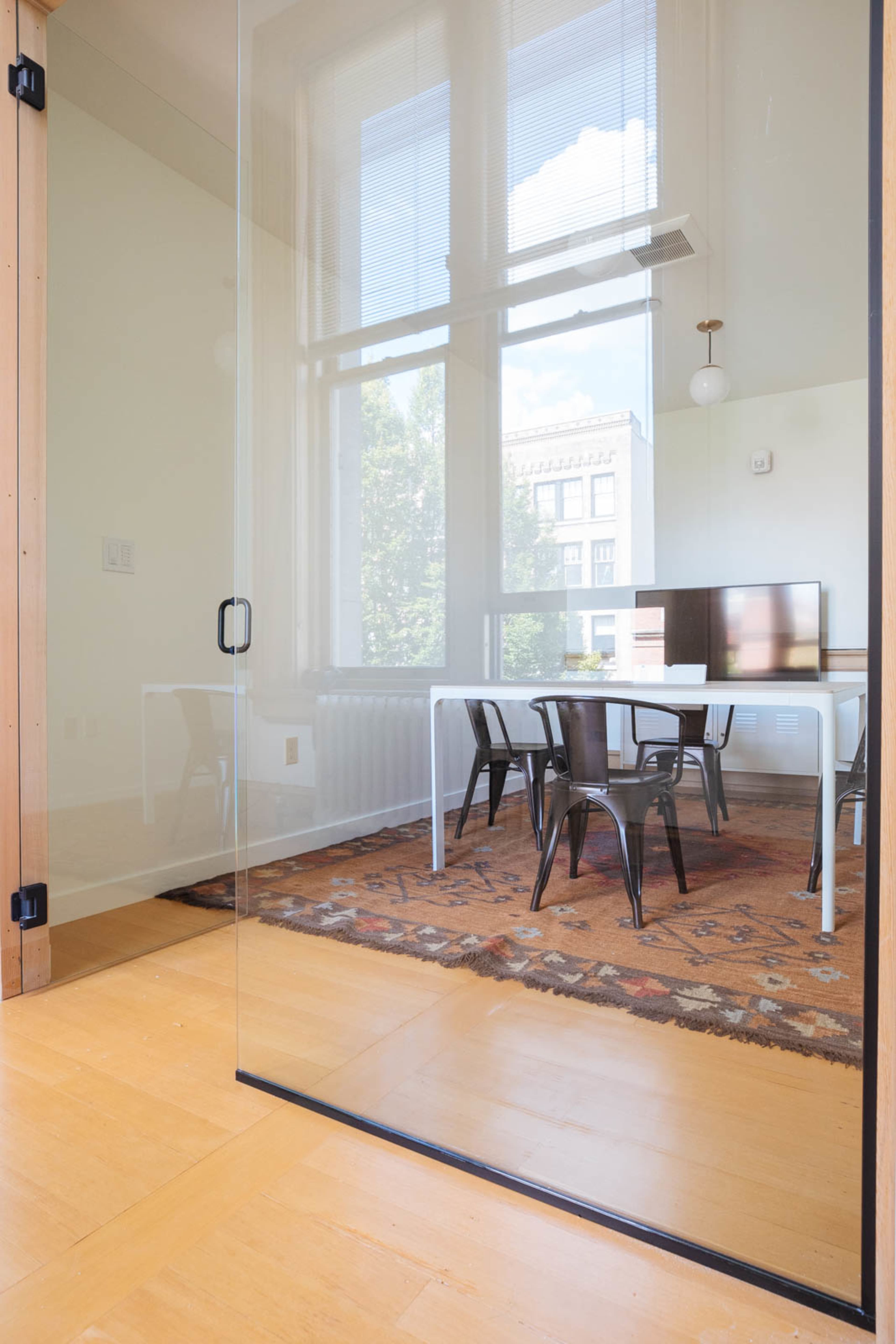 A glass wall separates a bright, minimalistic meeting room with a wooden table and metal chairs from an adjacent space.