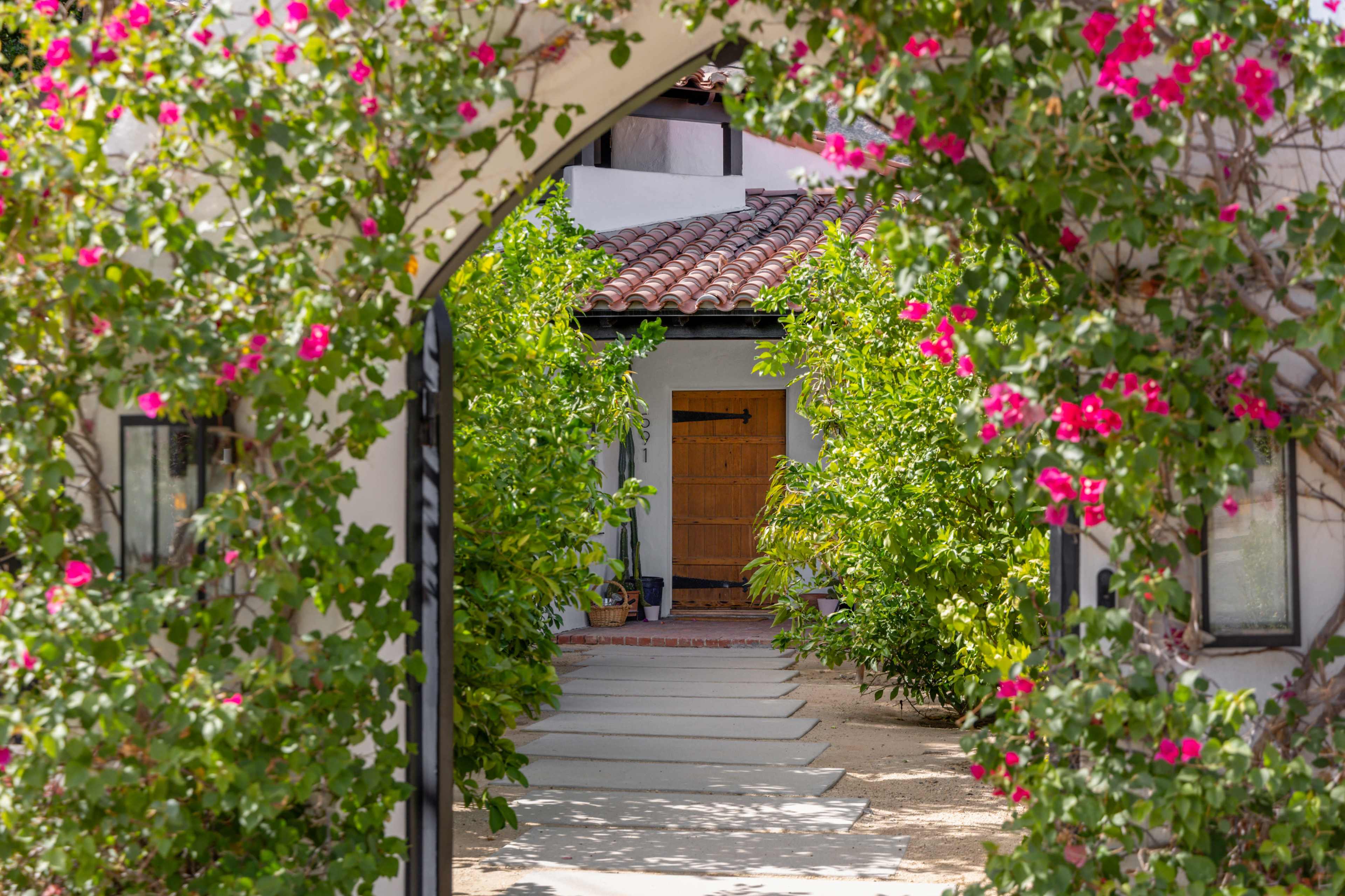 A wooden door is framed by an archway covered with vibrant pink flowers, leading to a pathway lined with greenery.