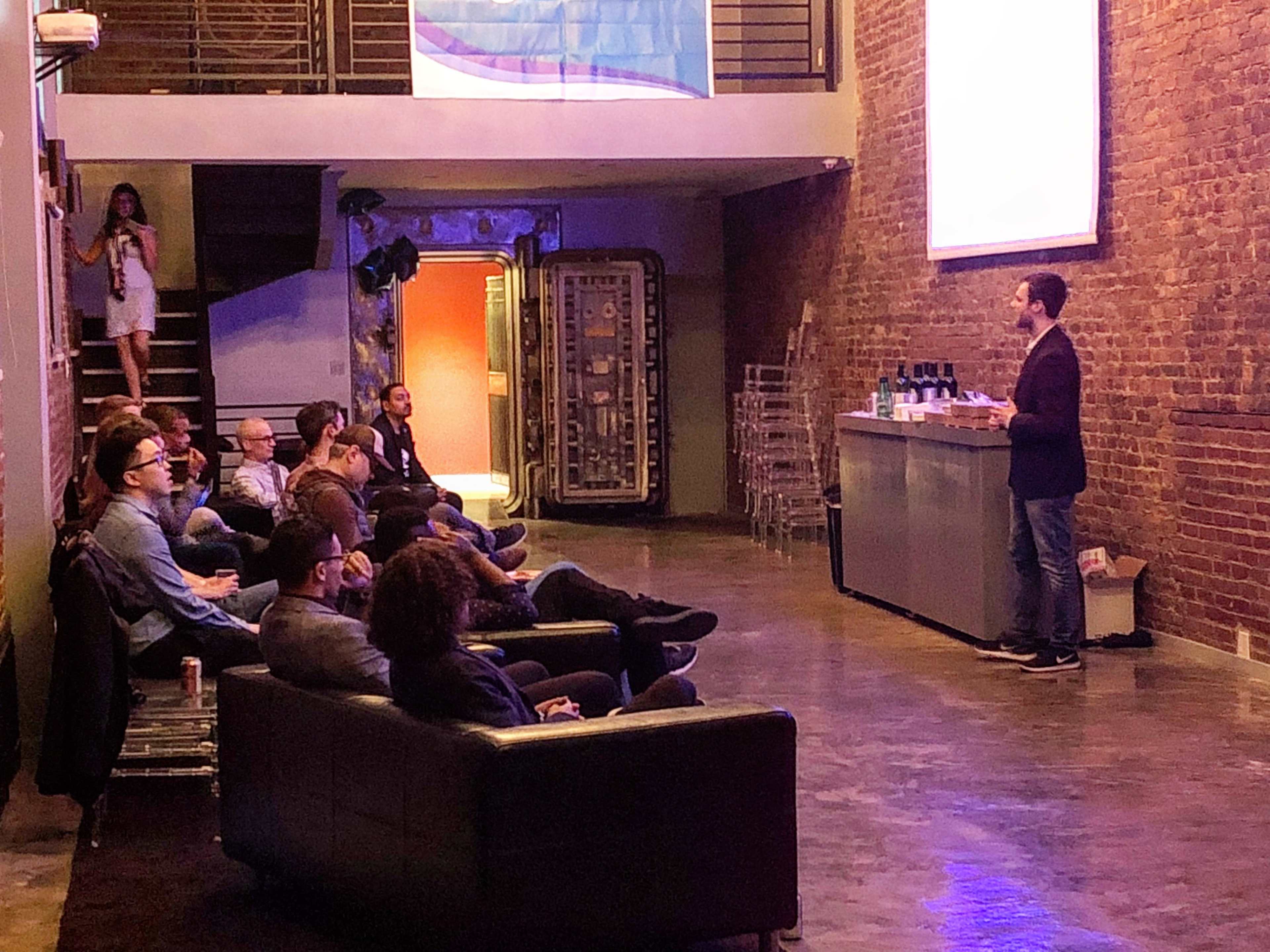A speaker stands in front of an audience in a dimly lit room with exposed brick walls and a bar area.
