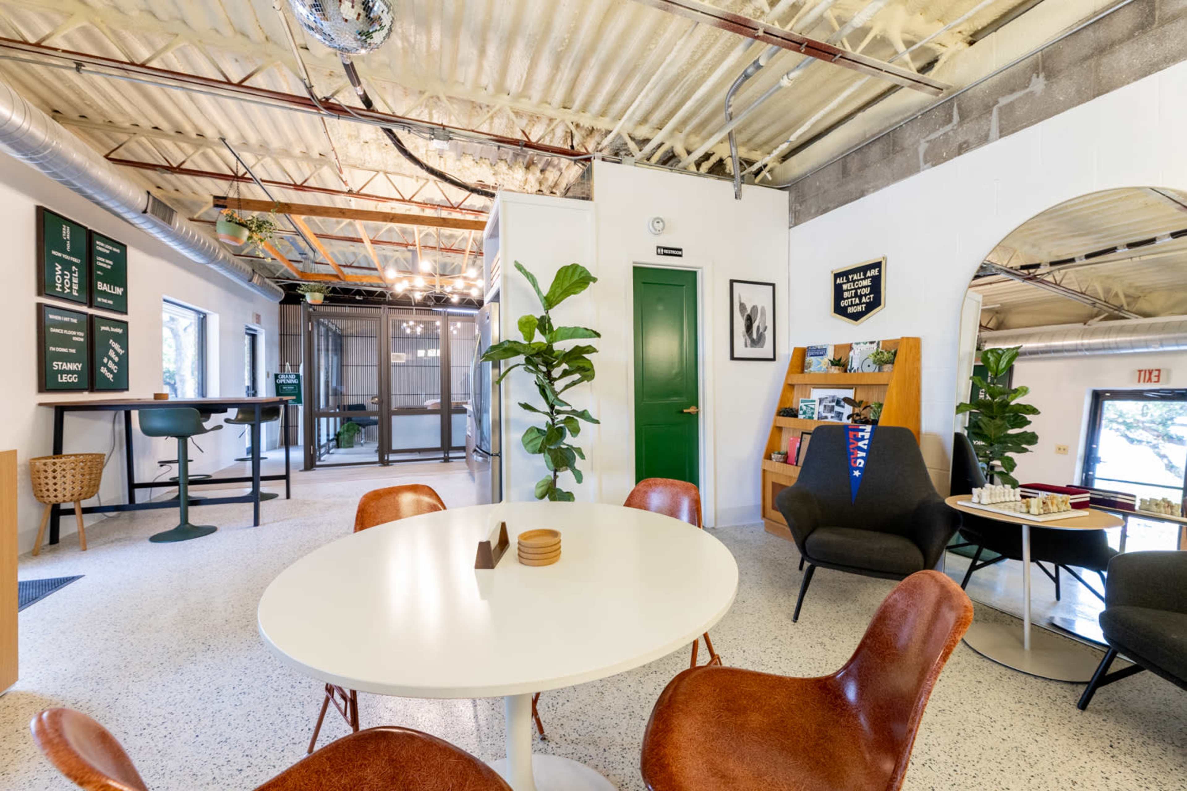 The image shows a modern, open-concept workspace featuring a round white table surrounded by brown chairs, with greenery and contemporary decor in the background.
