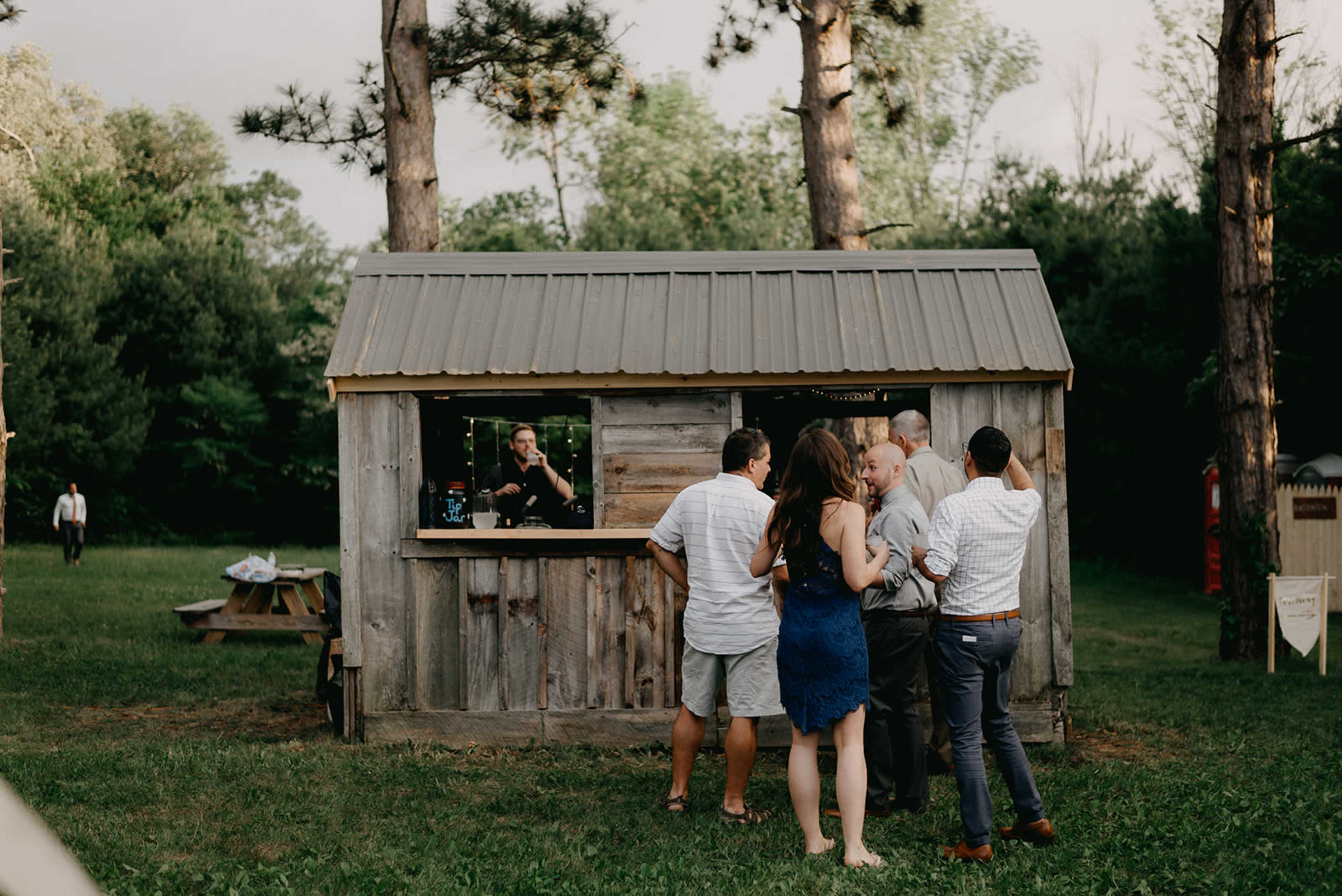 A group of people is gathered near a rustic bar with a corrugated metal roof surrounded by trees.