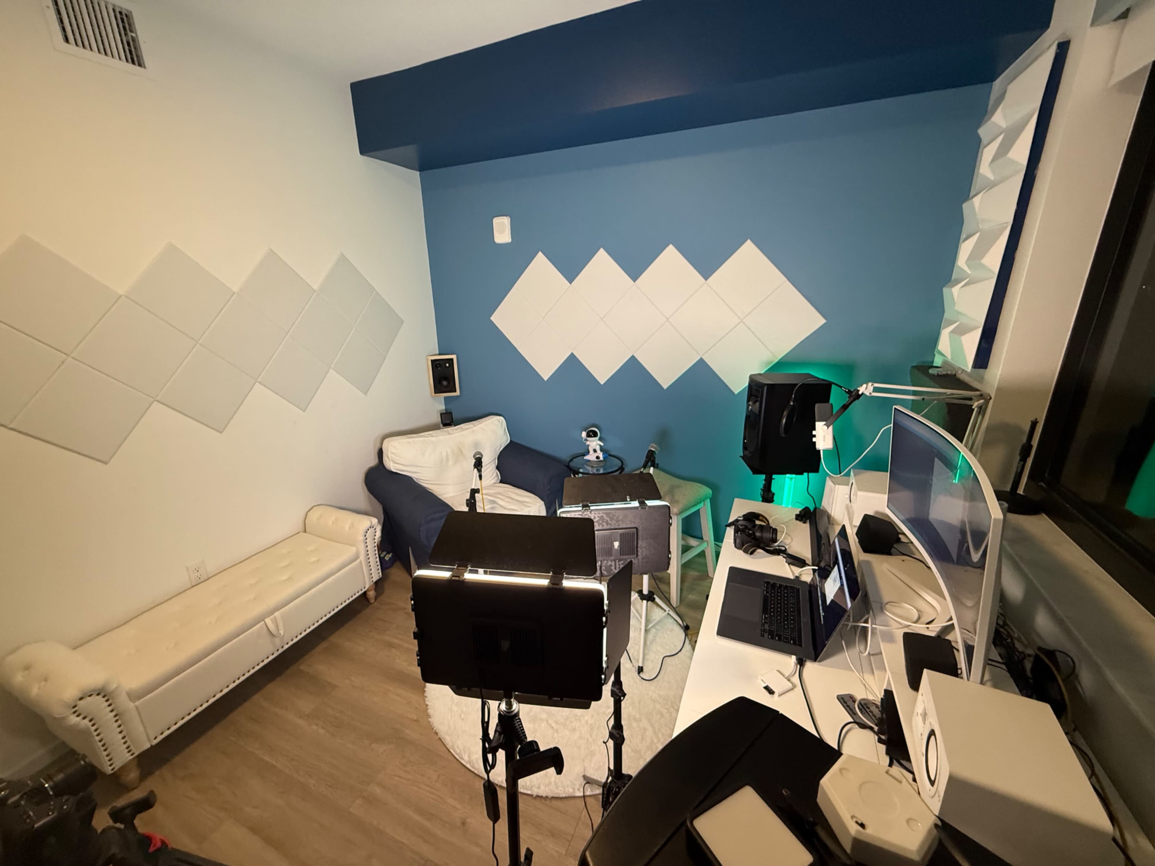 A well-organized room features a couch, a desk with a laptop, and various lighting equipment, set against a blue and white geometric wall design.