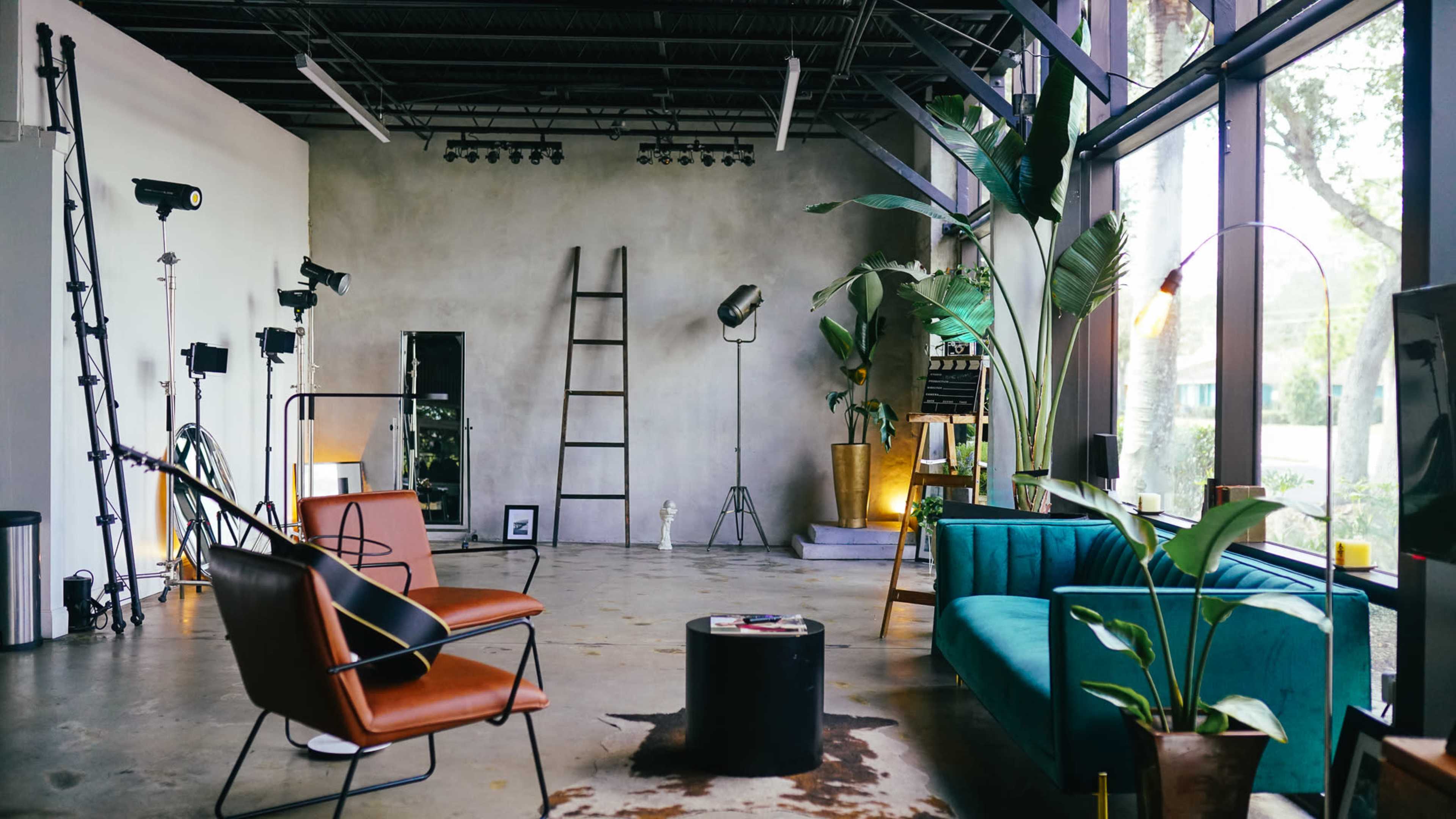 The image shows a spacious, well-lit studio with modern furniture, including a brown chair and a teal sofa, surrounded by plants and various lighting equipment.