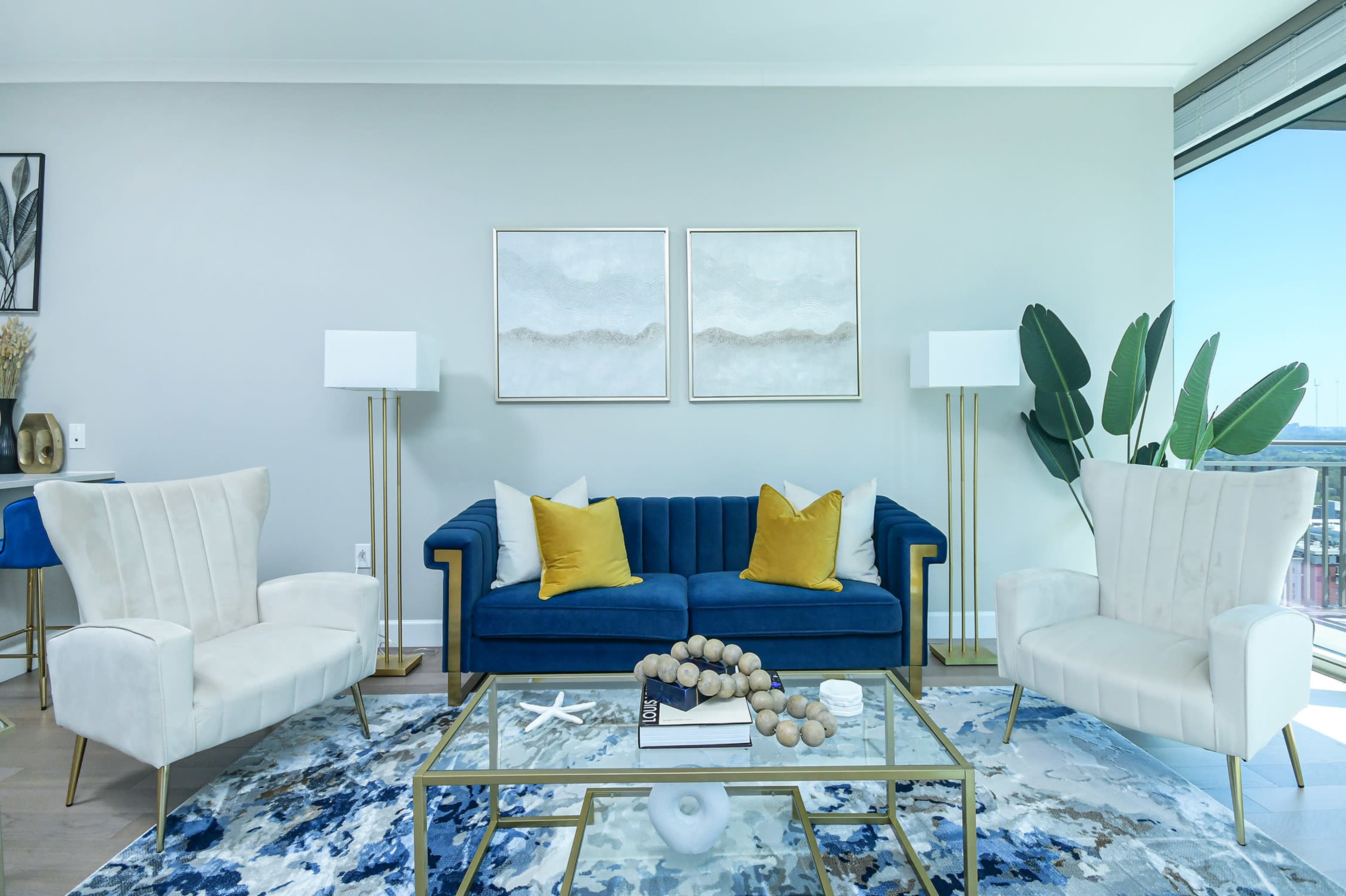 A modern living room featuring a blue sofa with yellow cushions, two white armchairs, a glass coffee table, and elegant artwork on the walls.