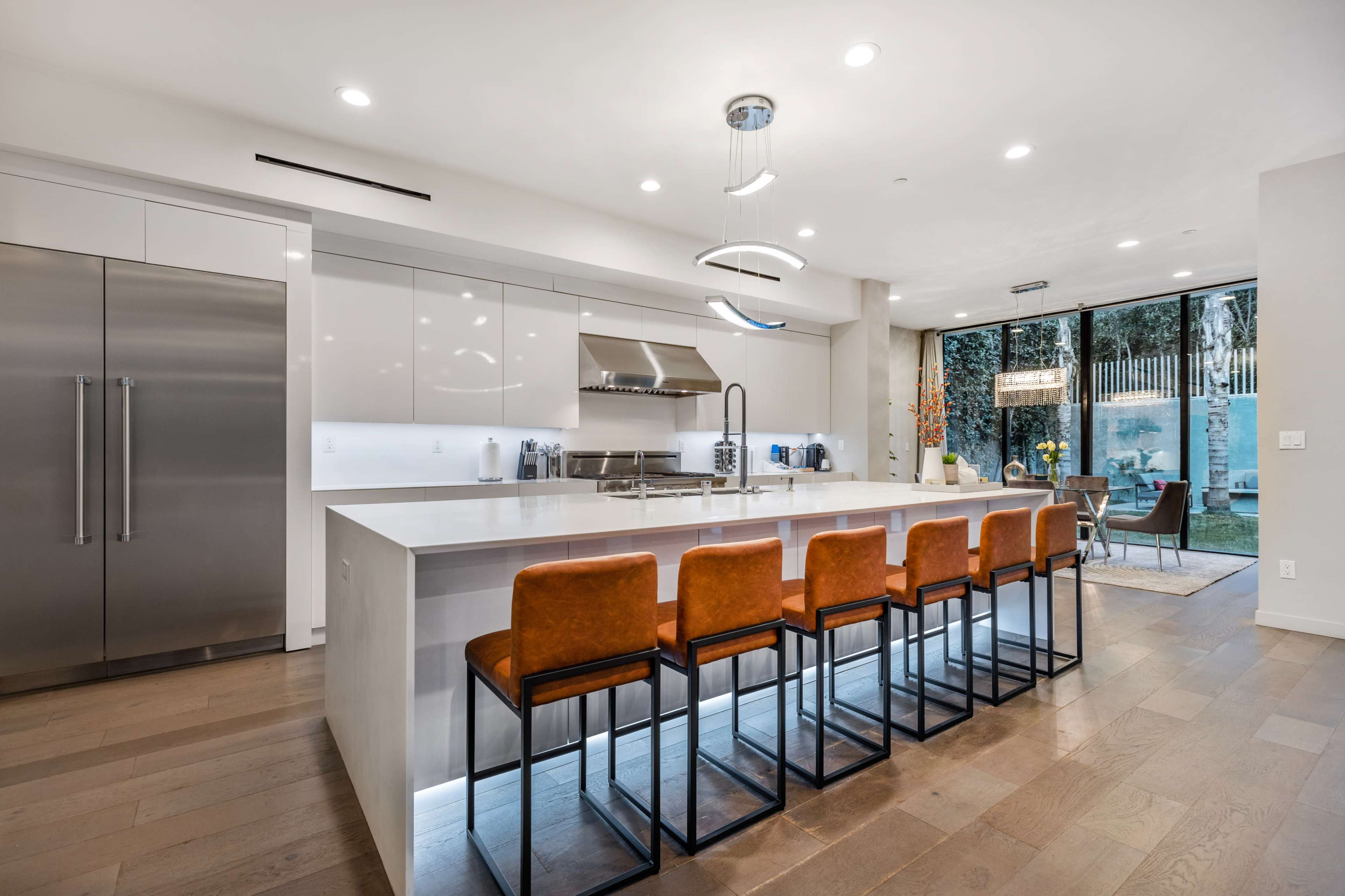 The kitchen features a modern design with white cabinetry, a large central island with orange bar stools, and stainless steel appliances, opening up to a backyard through glass doors.