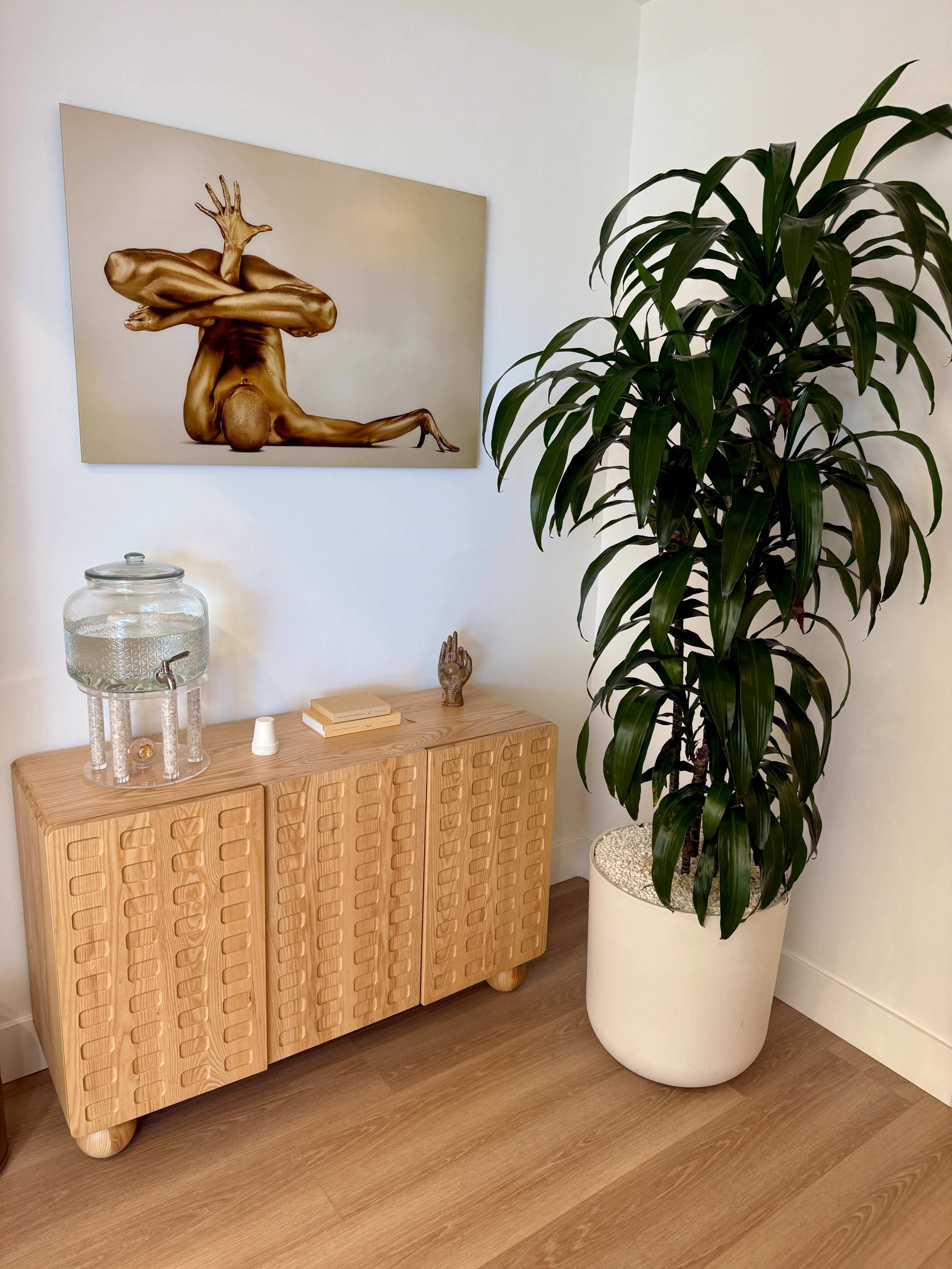 The image shows a wooden sideboard with textured details, a glass water dispenser on top, and a tall potted plant beside a wall with a painting of a posed figure.