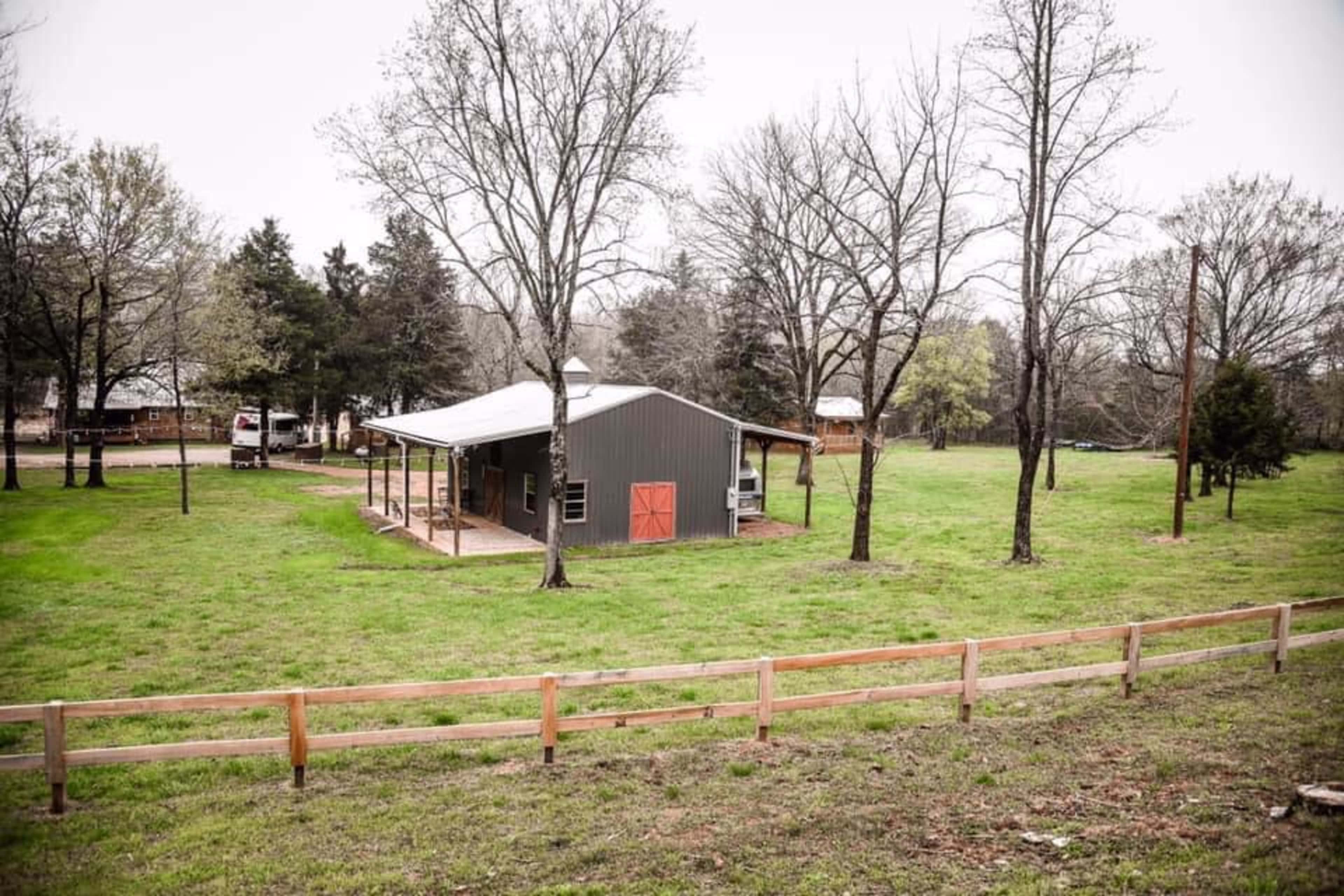A small gray house with a red door stands in a grassy area surrounded by bare trees and a wooden fence.