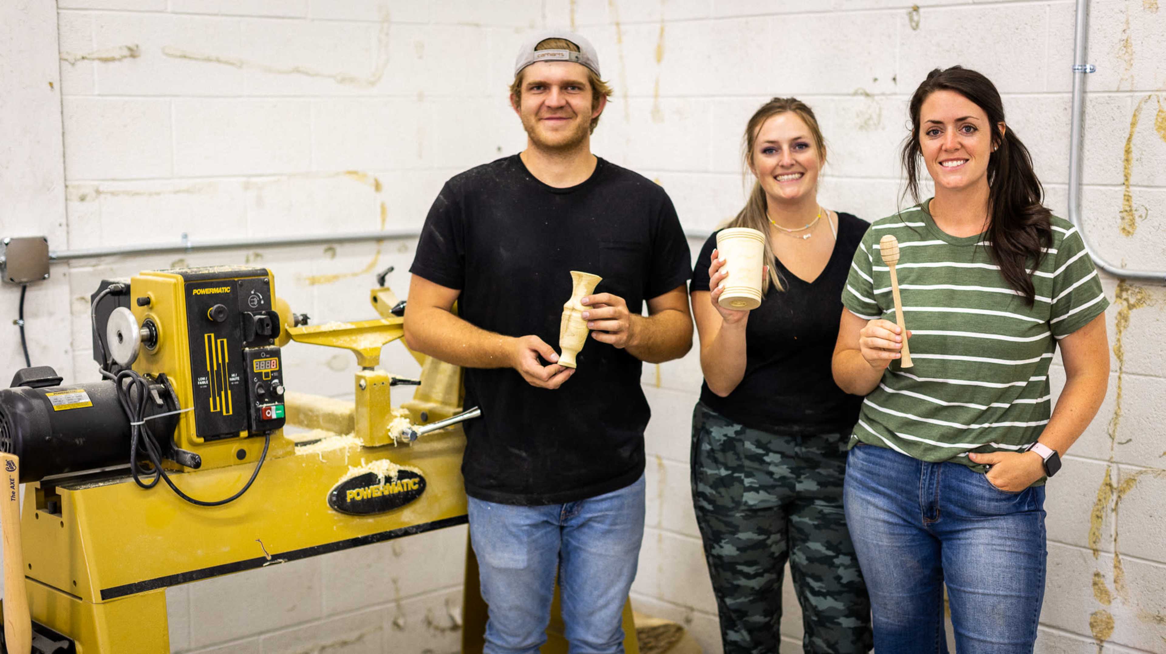 Three individuals stand in a workshop, holding wooden items and tools, with a lathe machine visible in the background.