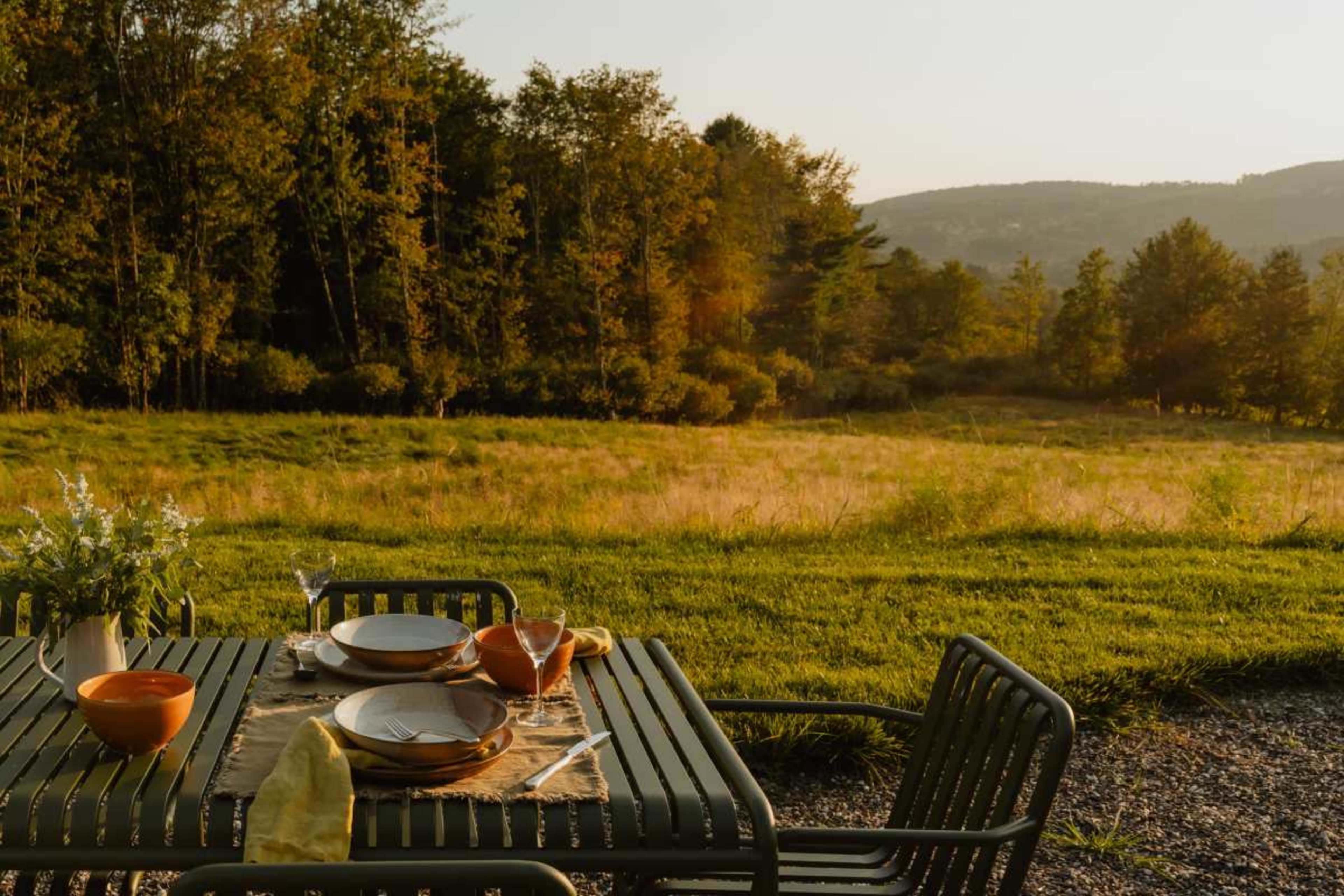 A dining table set with plates and cutlery overlooks a grassy field and wooded hills under a sunset sky.