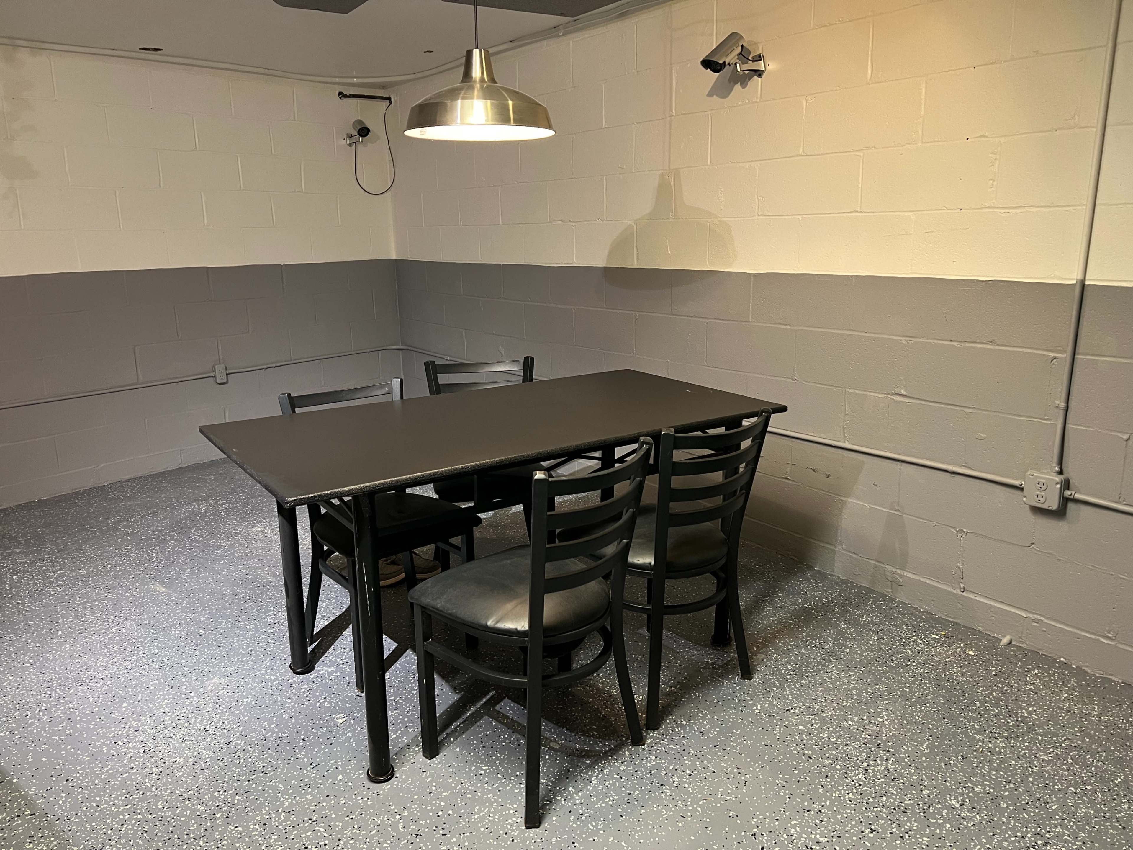 A black table with four chairs is positioned in a sparsely furnished room with gray walls and a ceiling-mounted lamp.