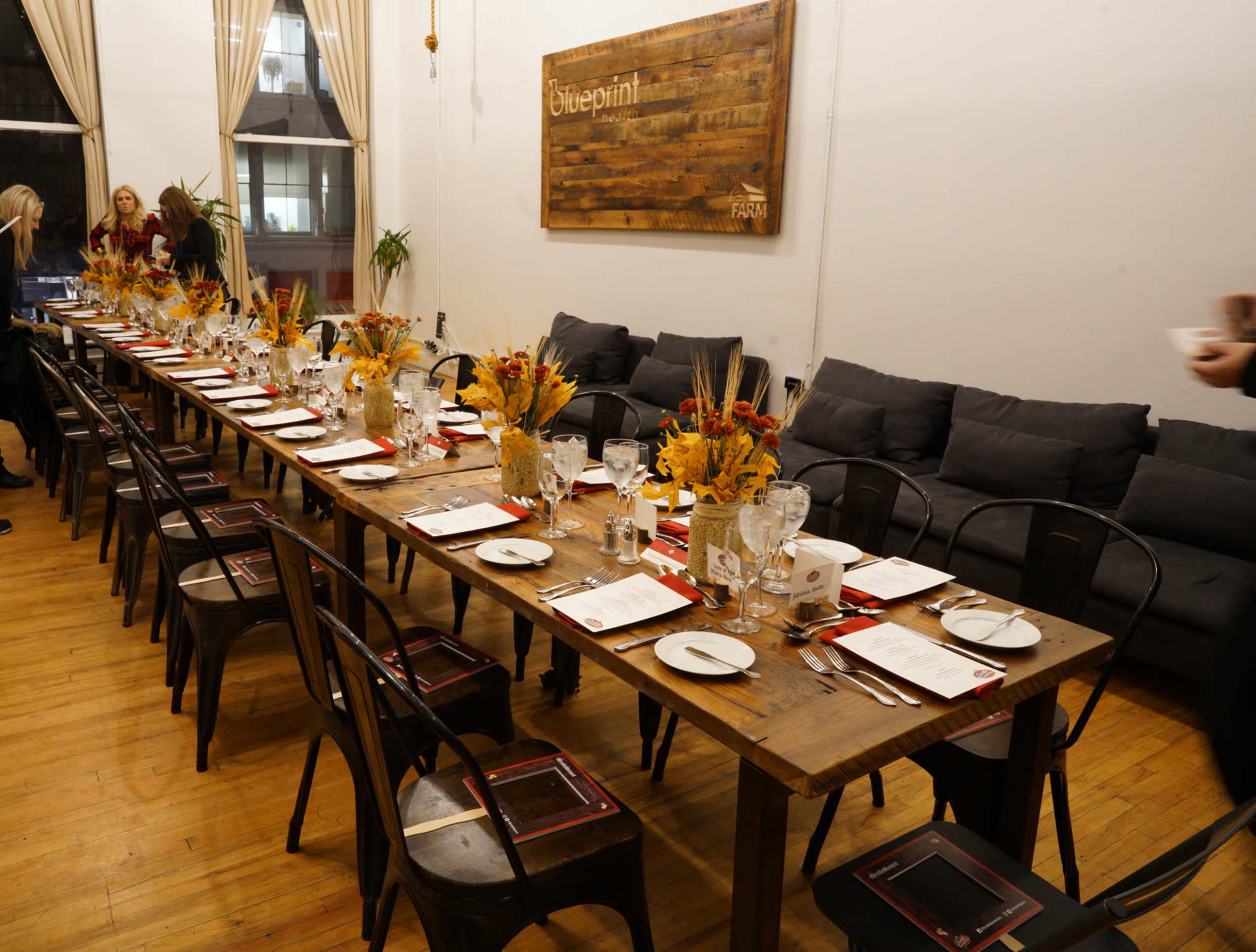 A long wooden dining table is set with plates and glasses, adorned with floral centerpieces, in a room featuring large windows and comfortable seating.