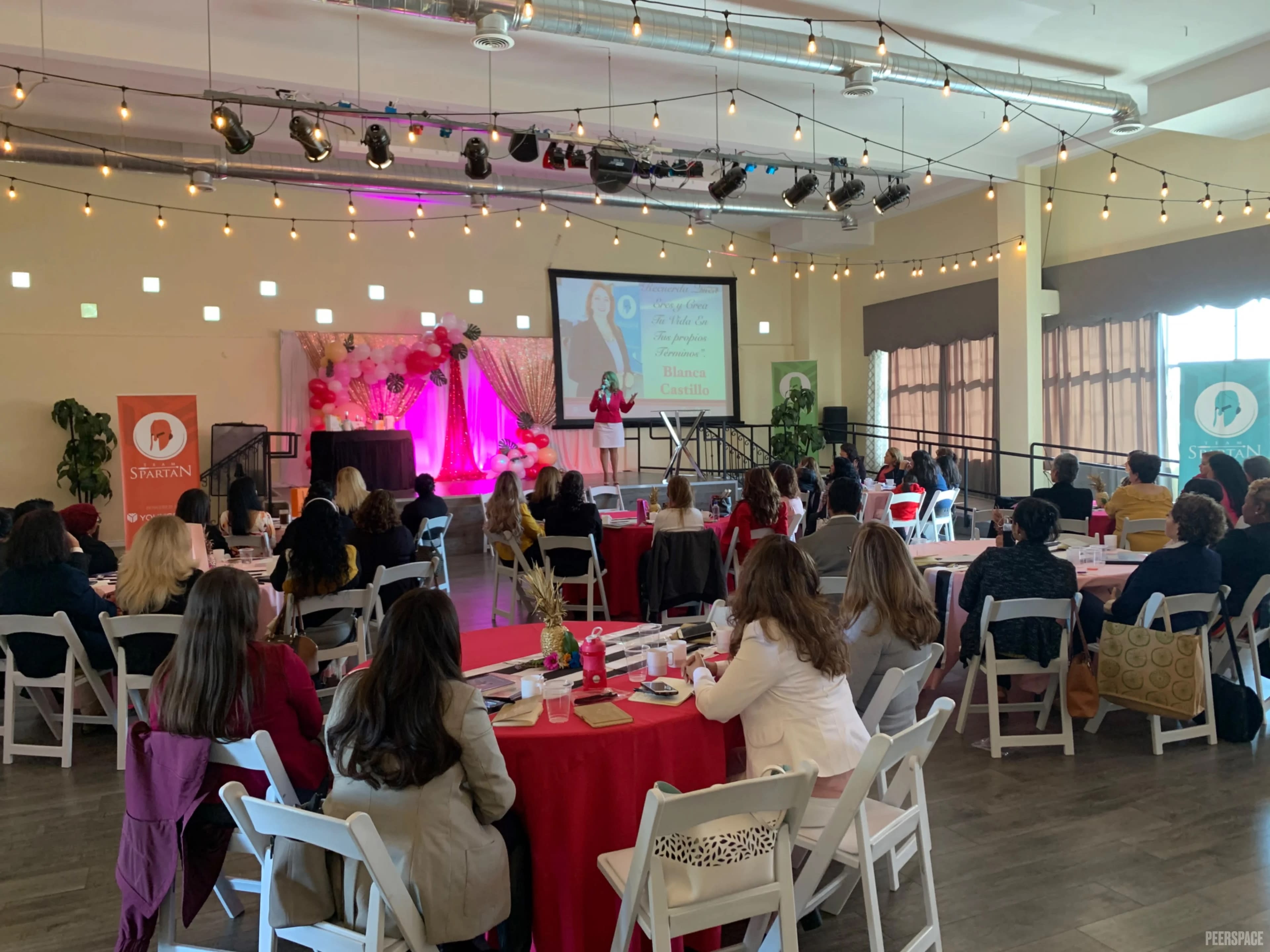 A speaker presents to an audience at a conference, with tables set for attendees and decorated in a vibrant, formal style.