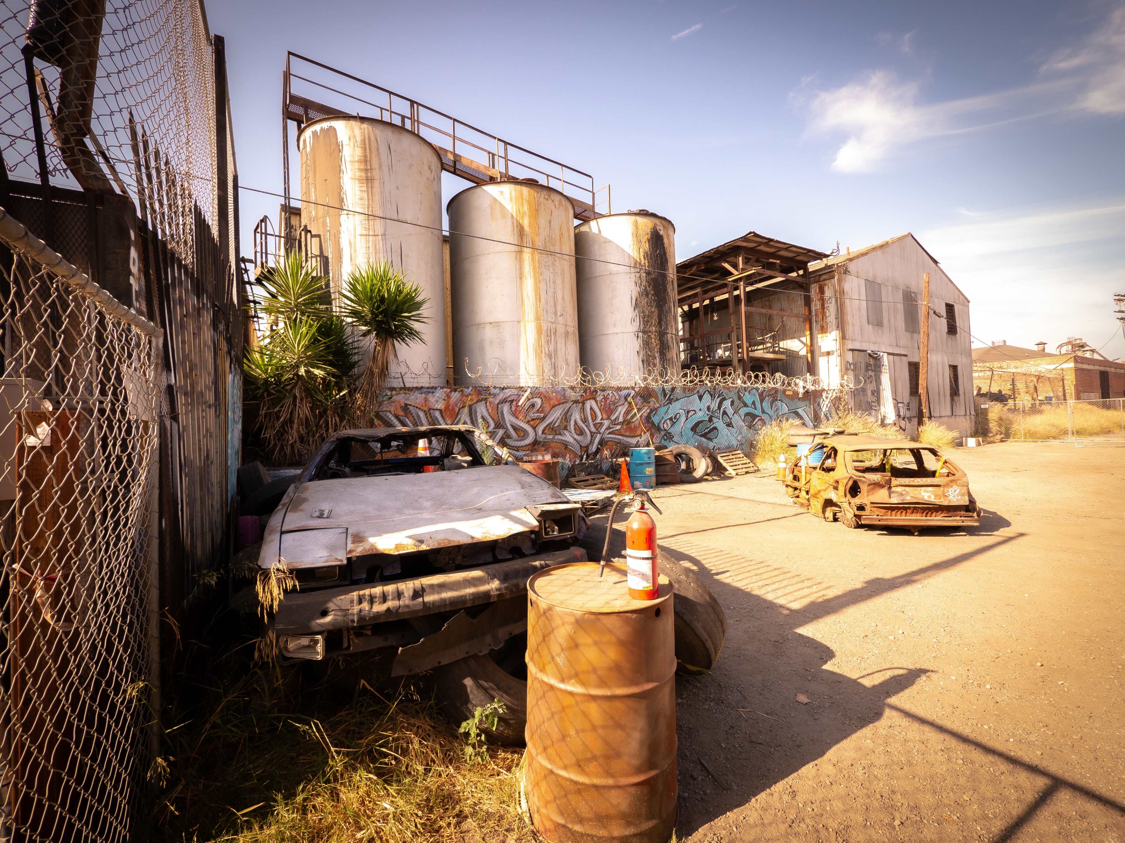 The image shows a deserted industrial site with rusted vehicles and large metal tanks in the background.