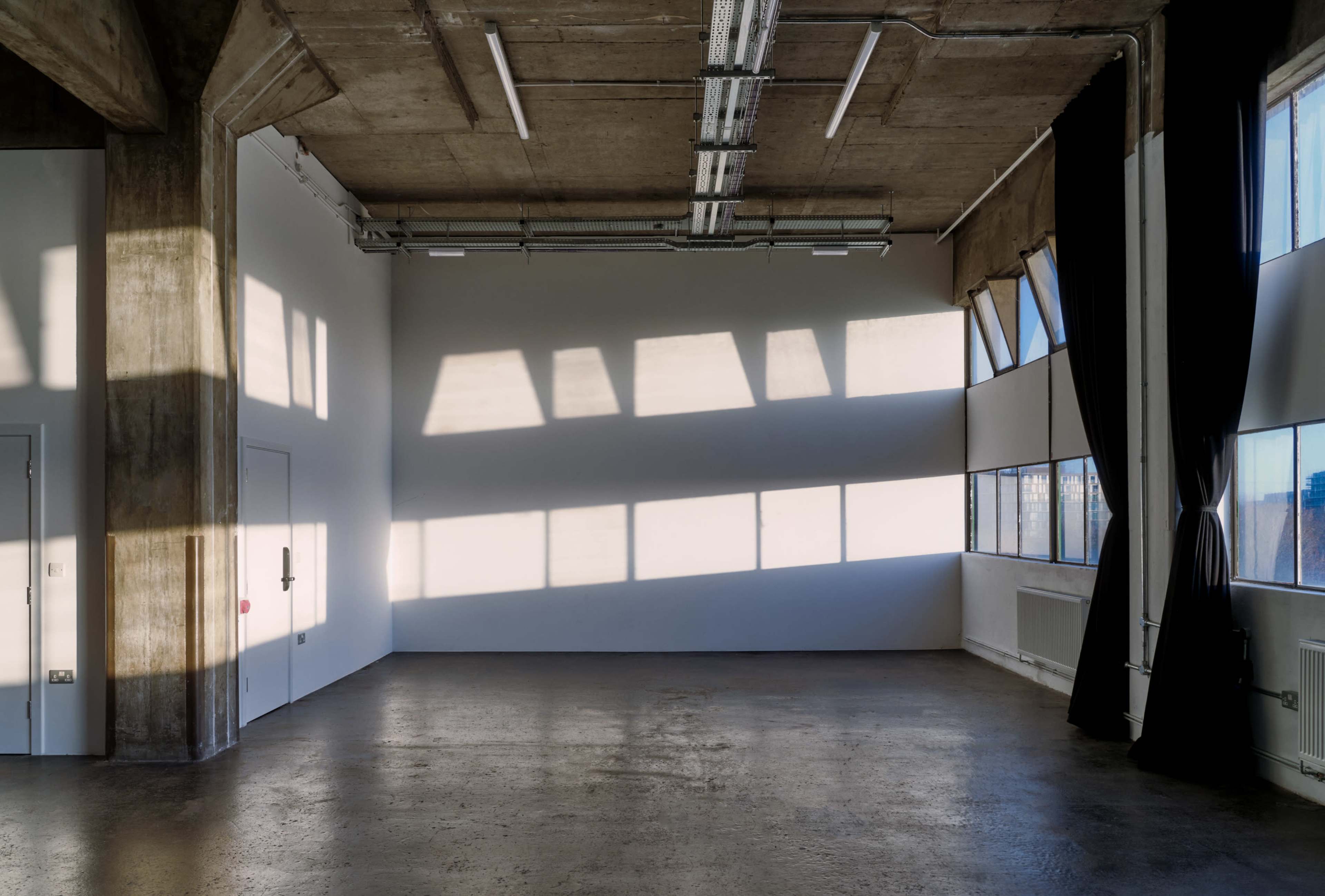 A spacious room with concrete walls and large windows casting shadows on the floor.