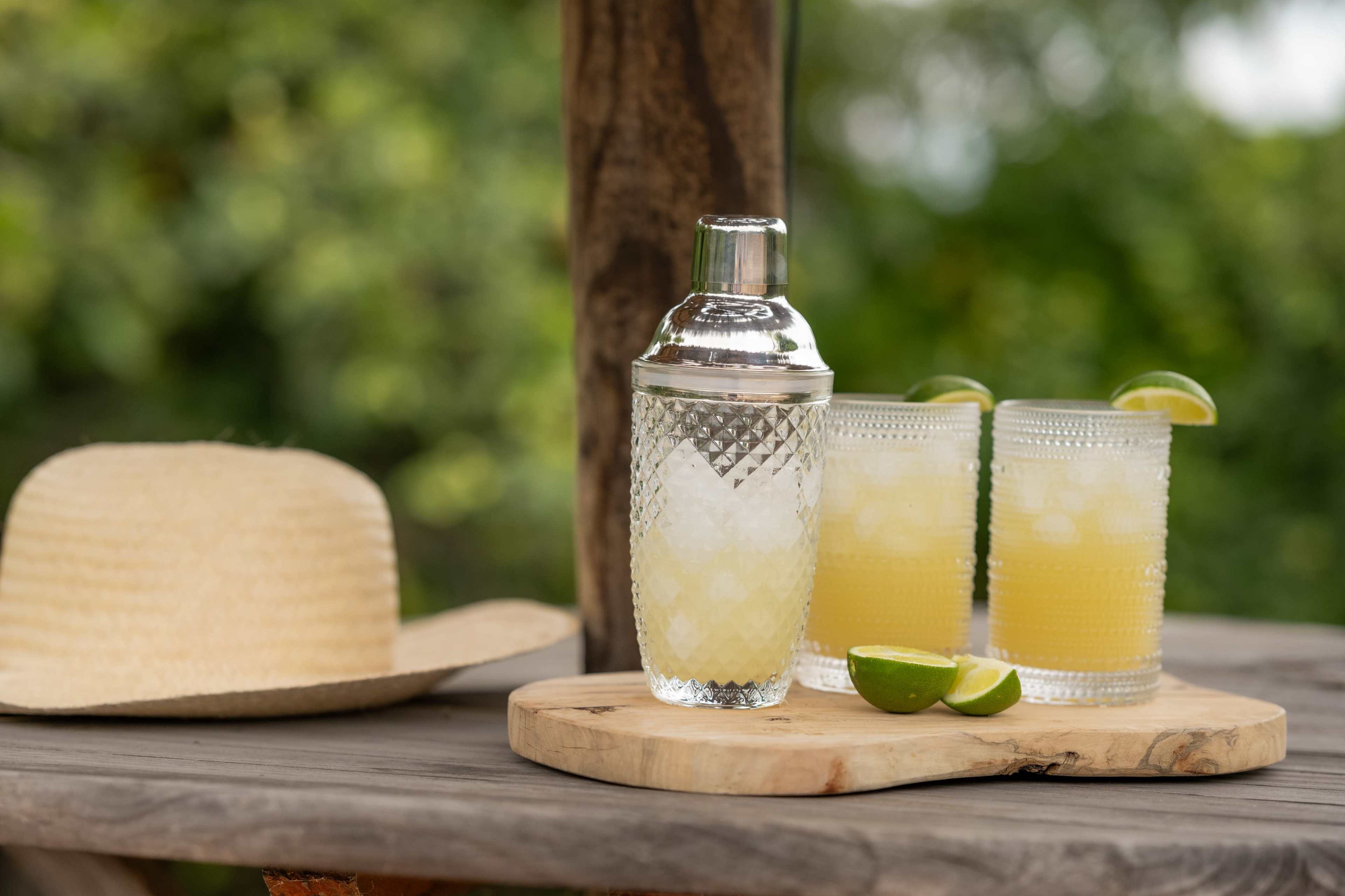 A cocktail shaker and two glasses of yellow drinks with lime wedges are arranged on a wooden table beside a straw hat.