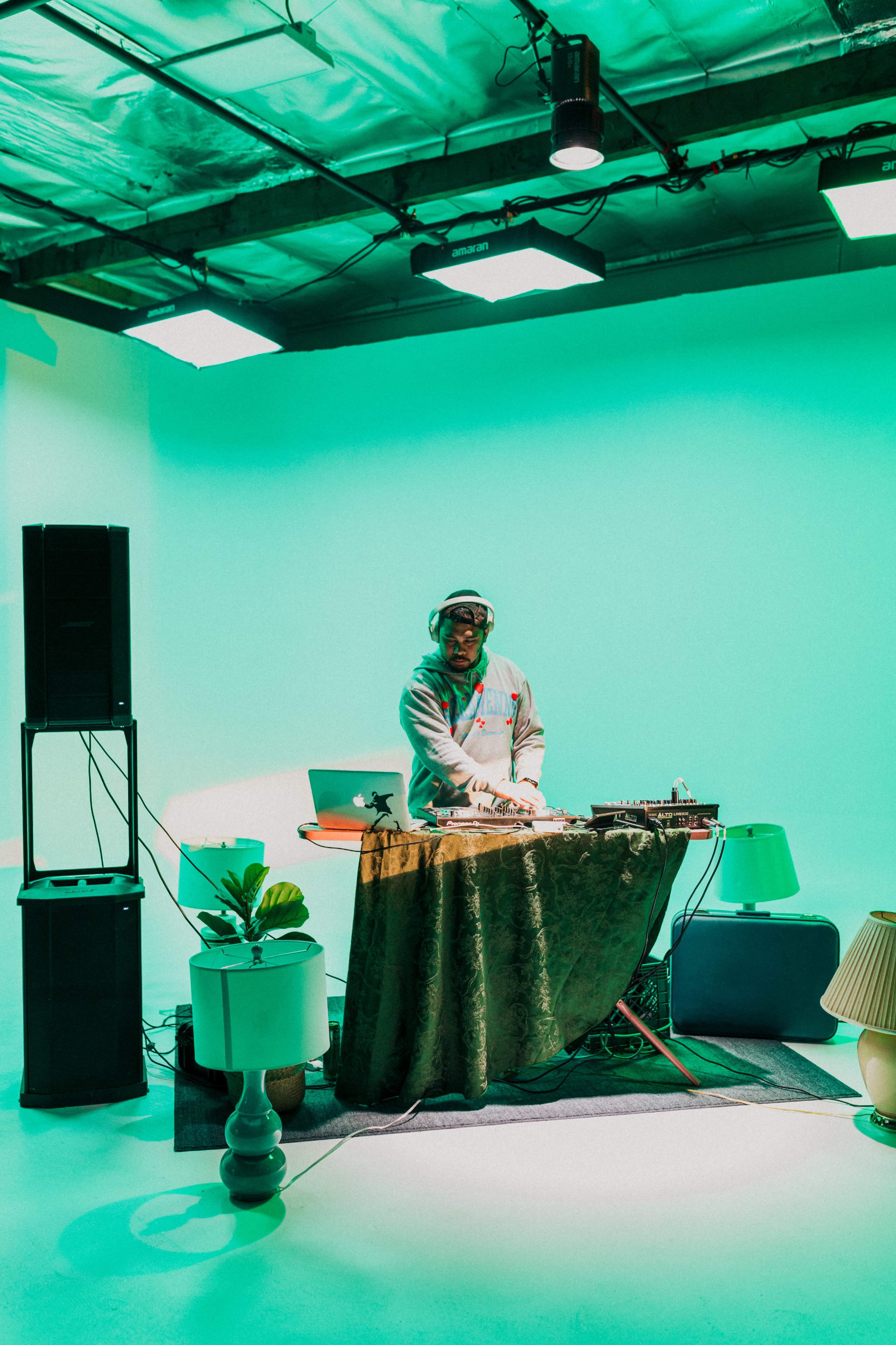 A DJ stands behind a table covered with a cloth, mixing music in a studio with green lighting and various decorative items around him.