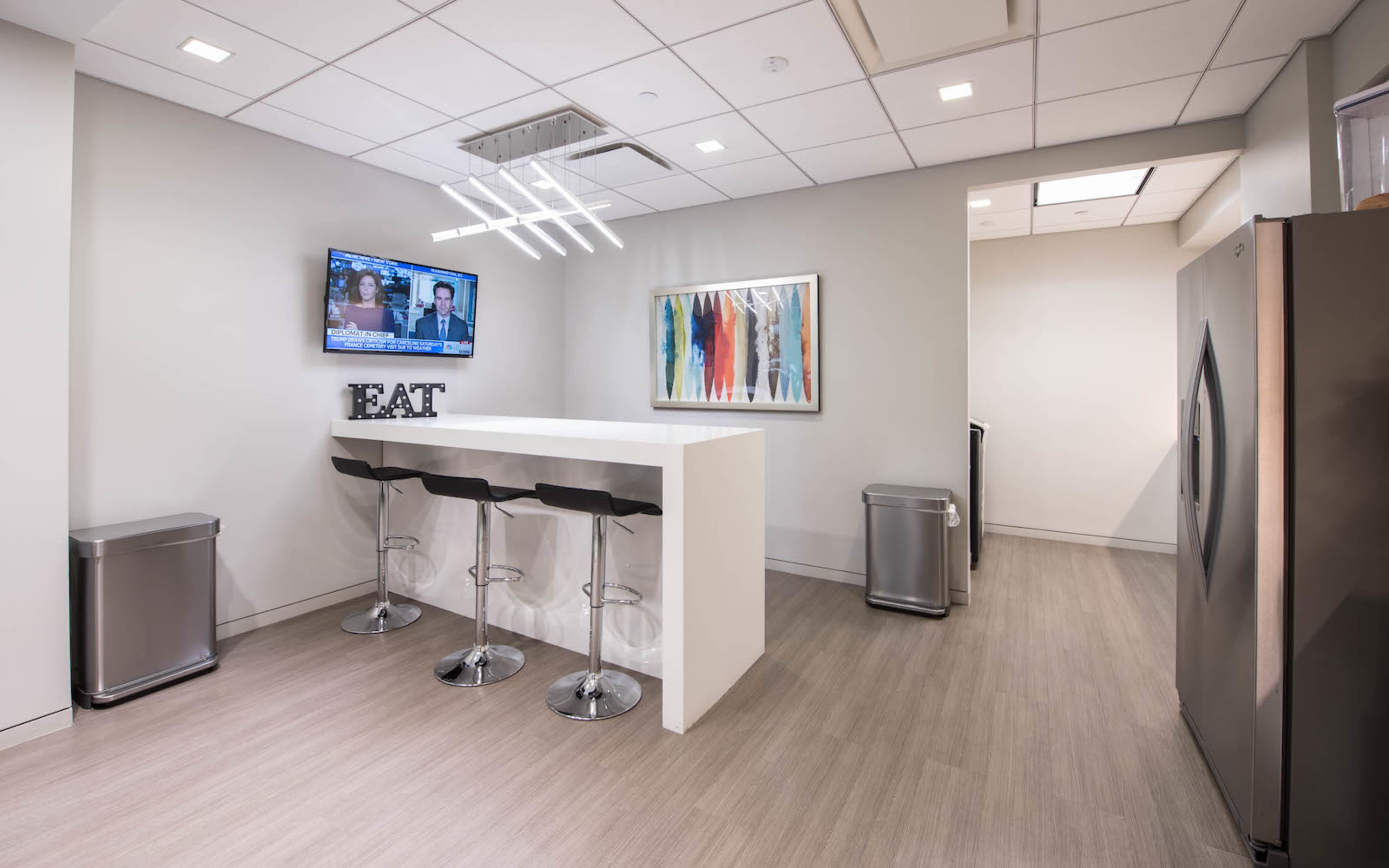 A modern kitchen area features a bar with three stools, a television mounted on the wall, and a stainless steel refrigerator.