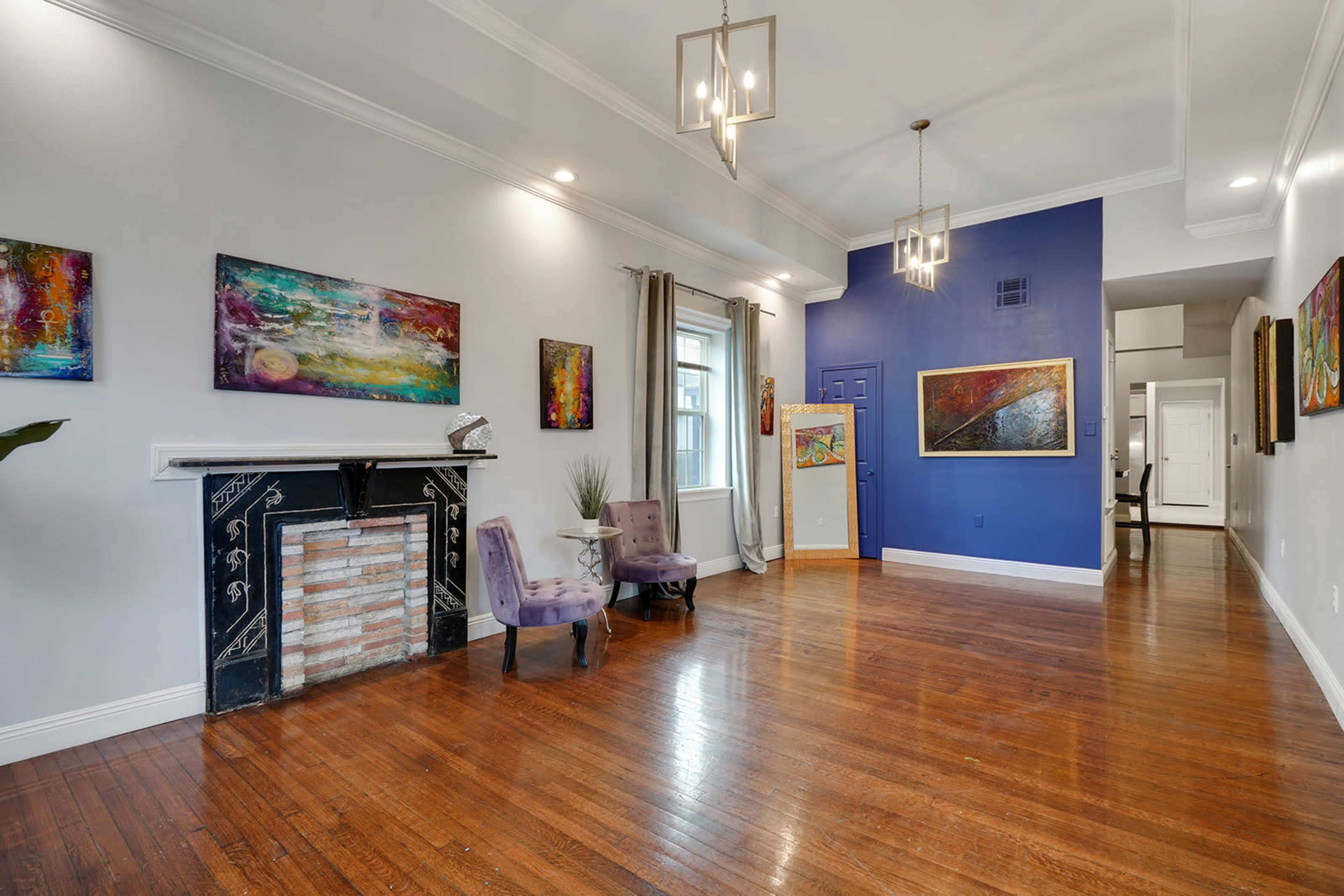 The image shows a spacious living room featuring wooden floors, a decorative fireplace, vibrant artwork on the walls, and large windows allowing light to enter the area.