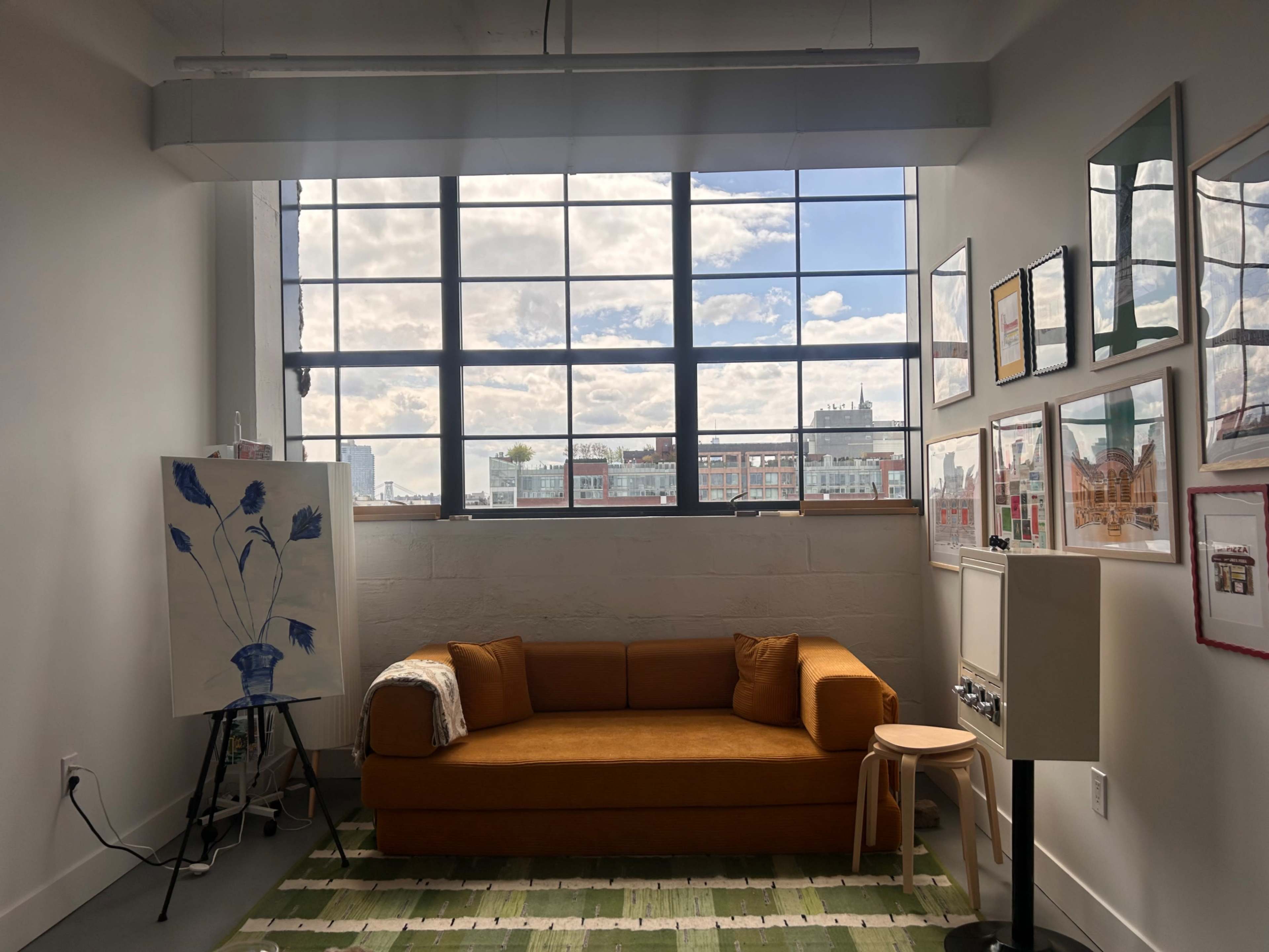 A bright room features a large window with a view of the sky, an orange couch, an easel with a painting, and framed artwork on the walls.