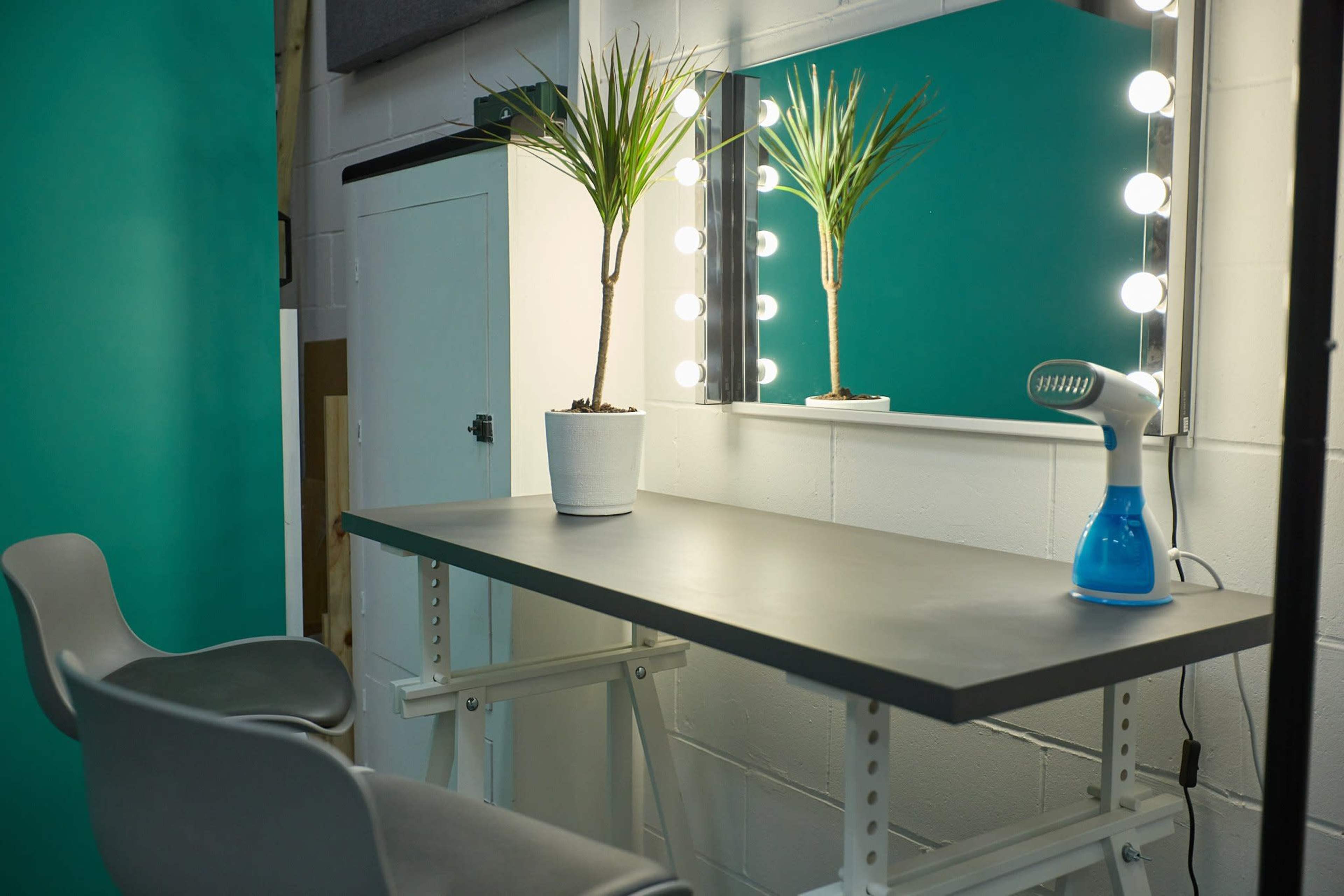 A minimalist dressing table with a mirror and bright lights, accompanied by two potted plants and a handheld steamer, is set against a teal wall.
