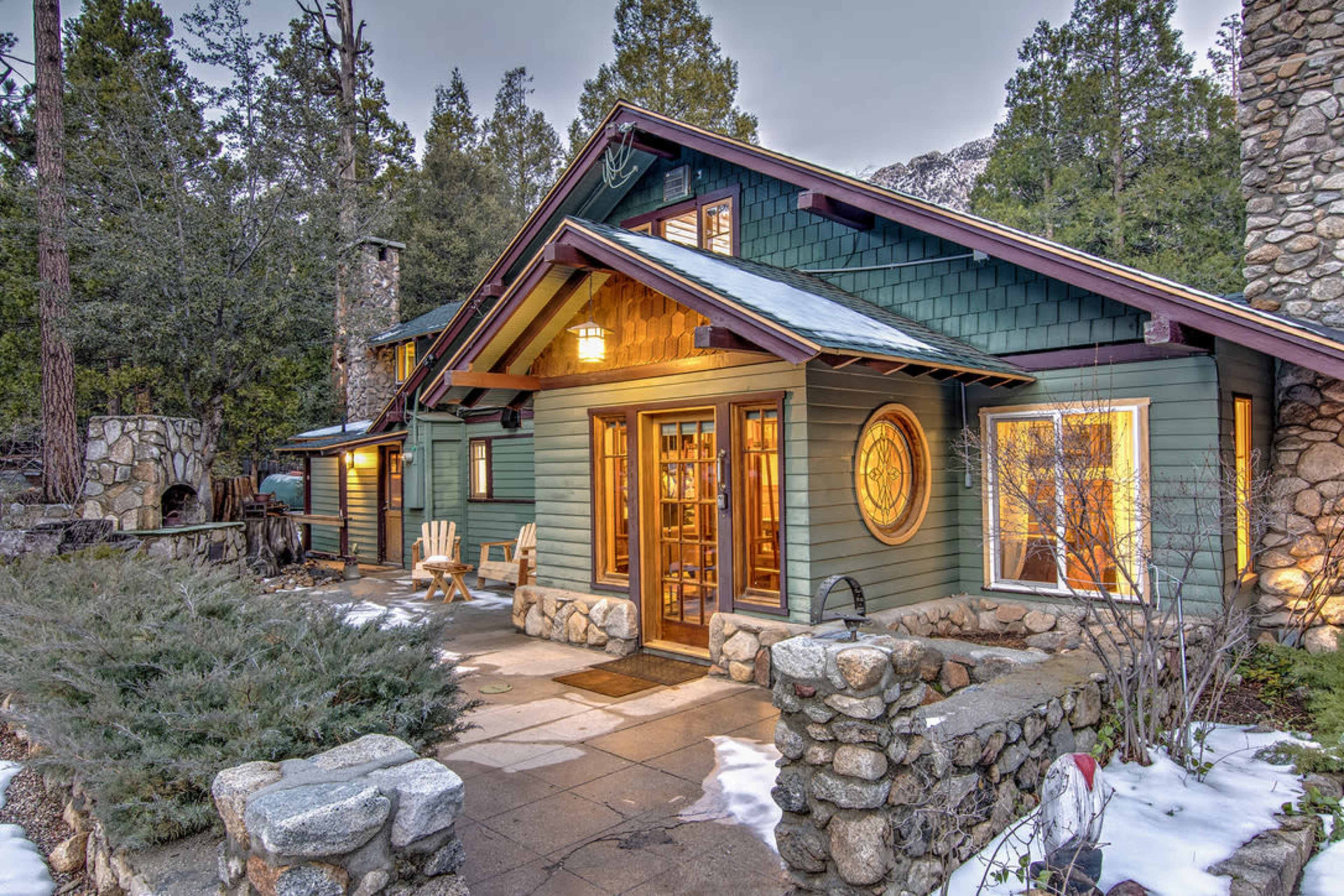 A stone and wood cabin with large windows and a welcoming porch is surrounded by evergreen trees and patches of snow.