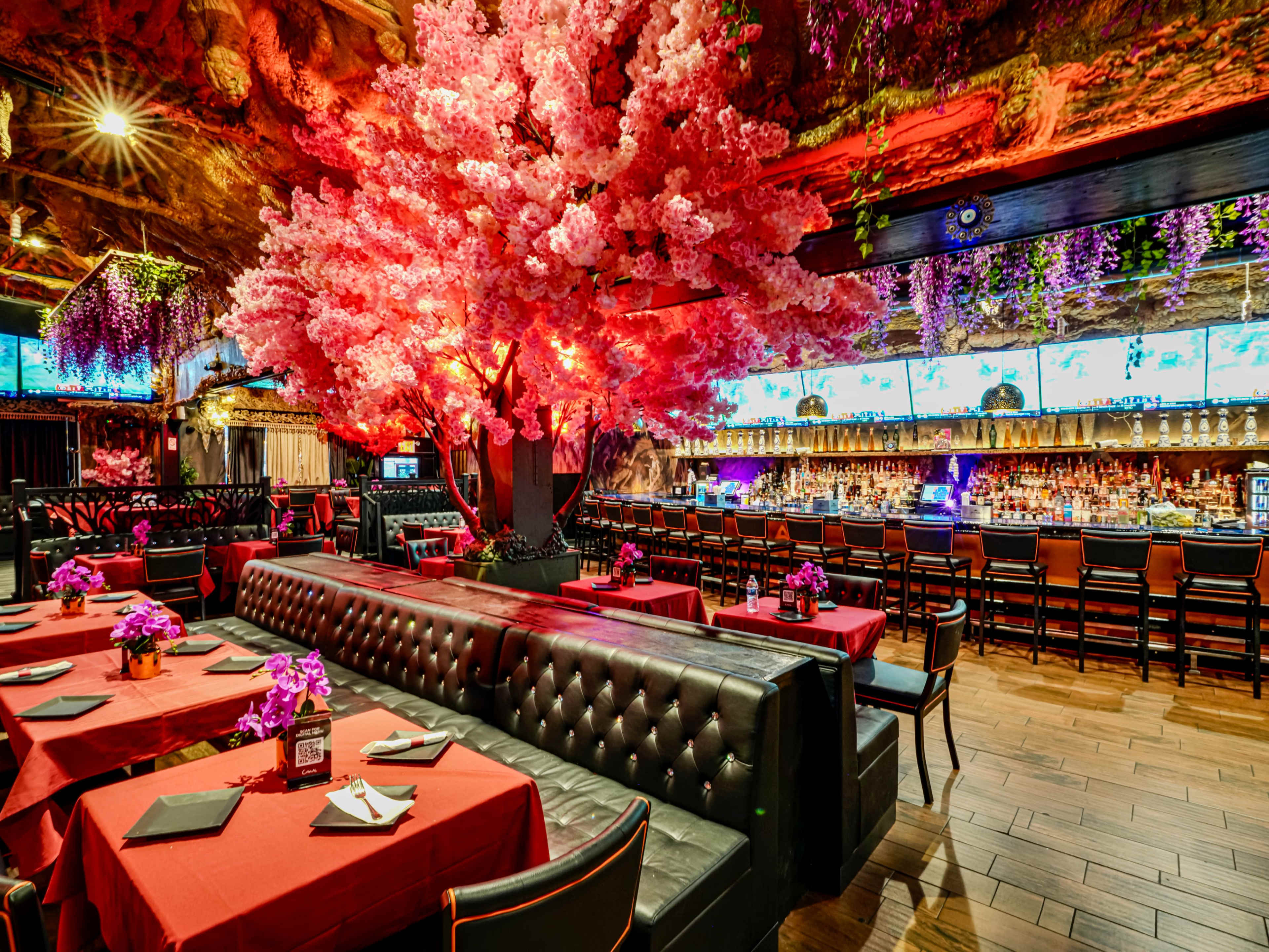 The image depicts a vibrant restaurant interior featuring a pink cherry blossom tree, red tablecloths, and a well-stocked bar in the background illuminated by soft lighting.