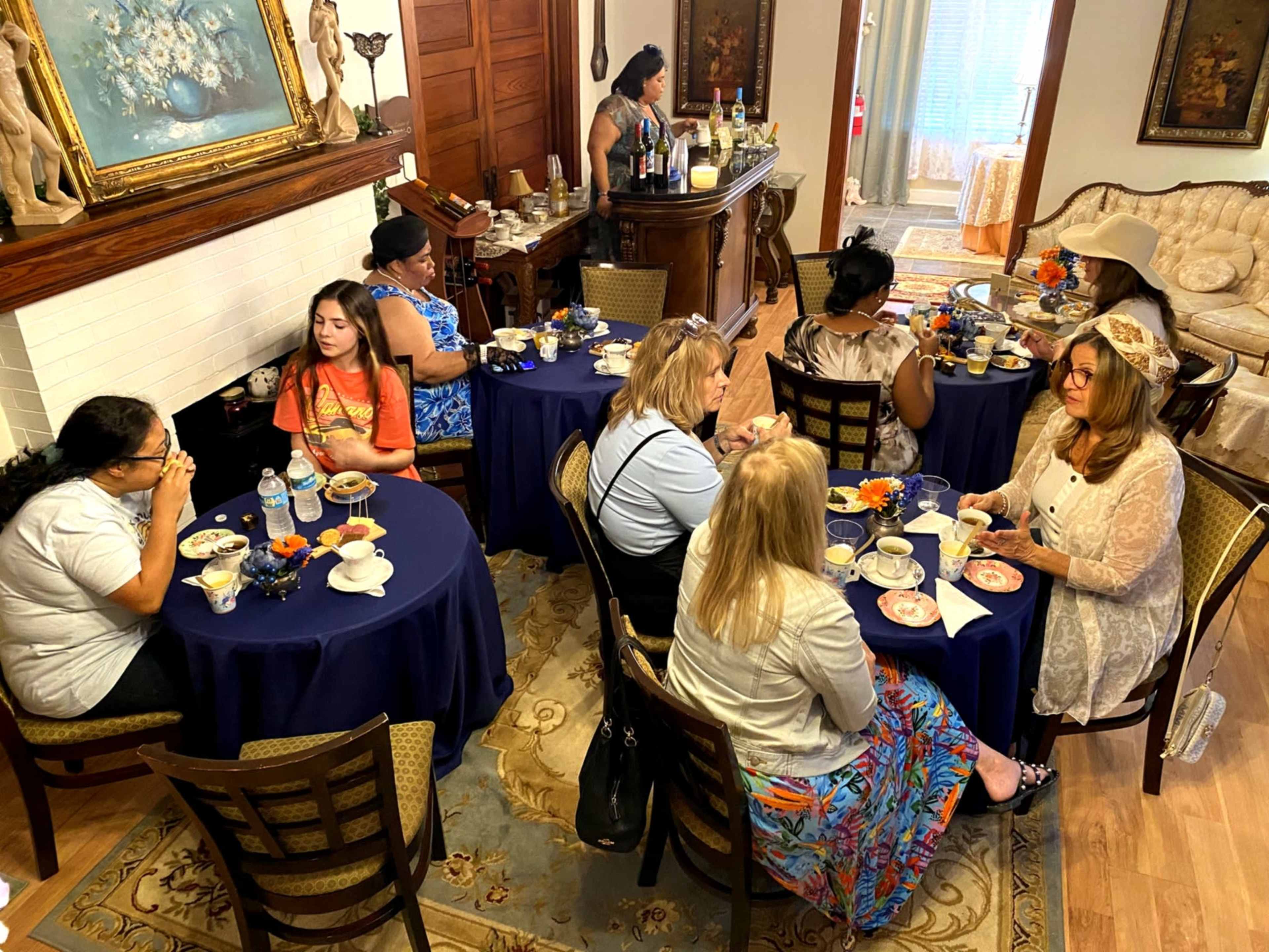 A group of women in a living room is enjoying a tea party at round tables with decorative table settings and various refreshments.