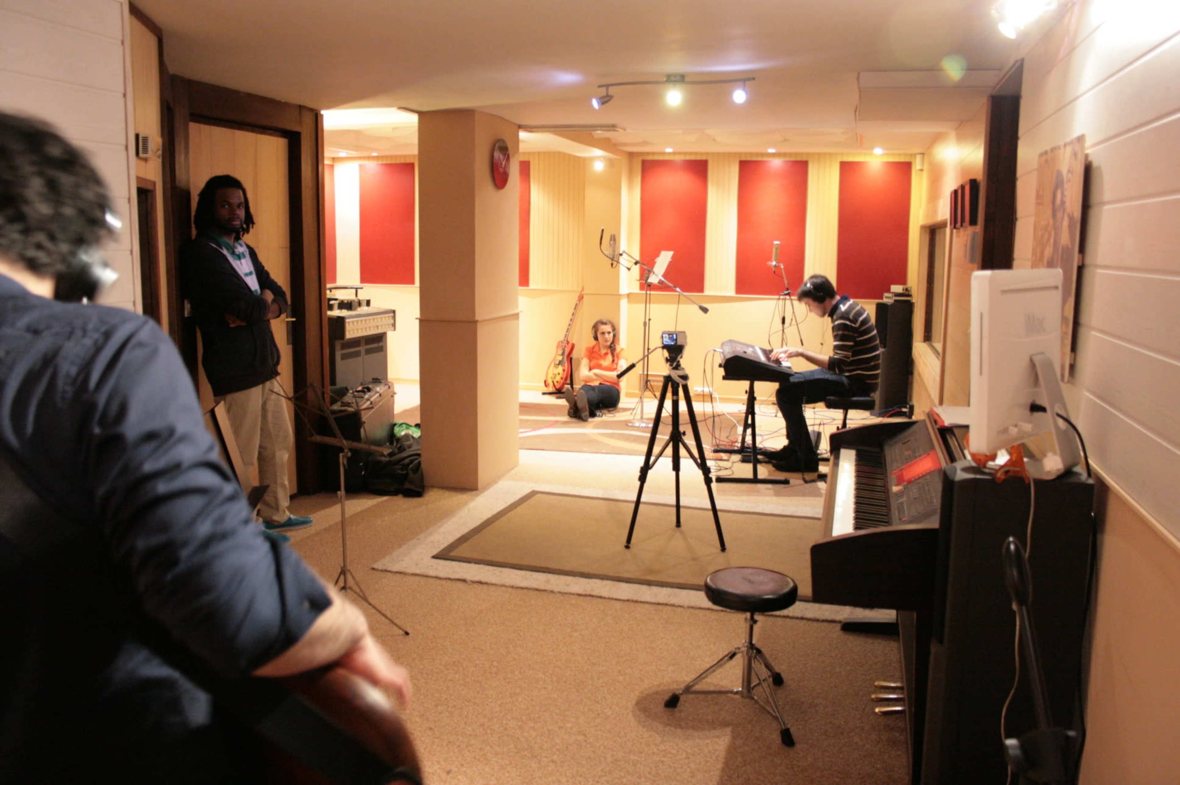 Musicians are seen rehearsing in a soundproof studio, with one playing guitar and another seated at a piano, while a microphone and cameras are set up for recording.