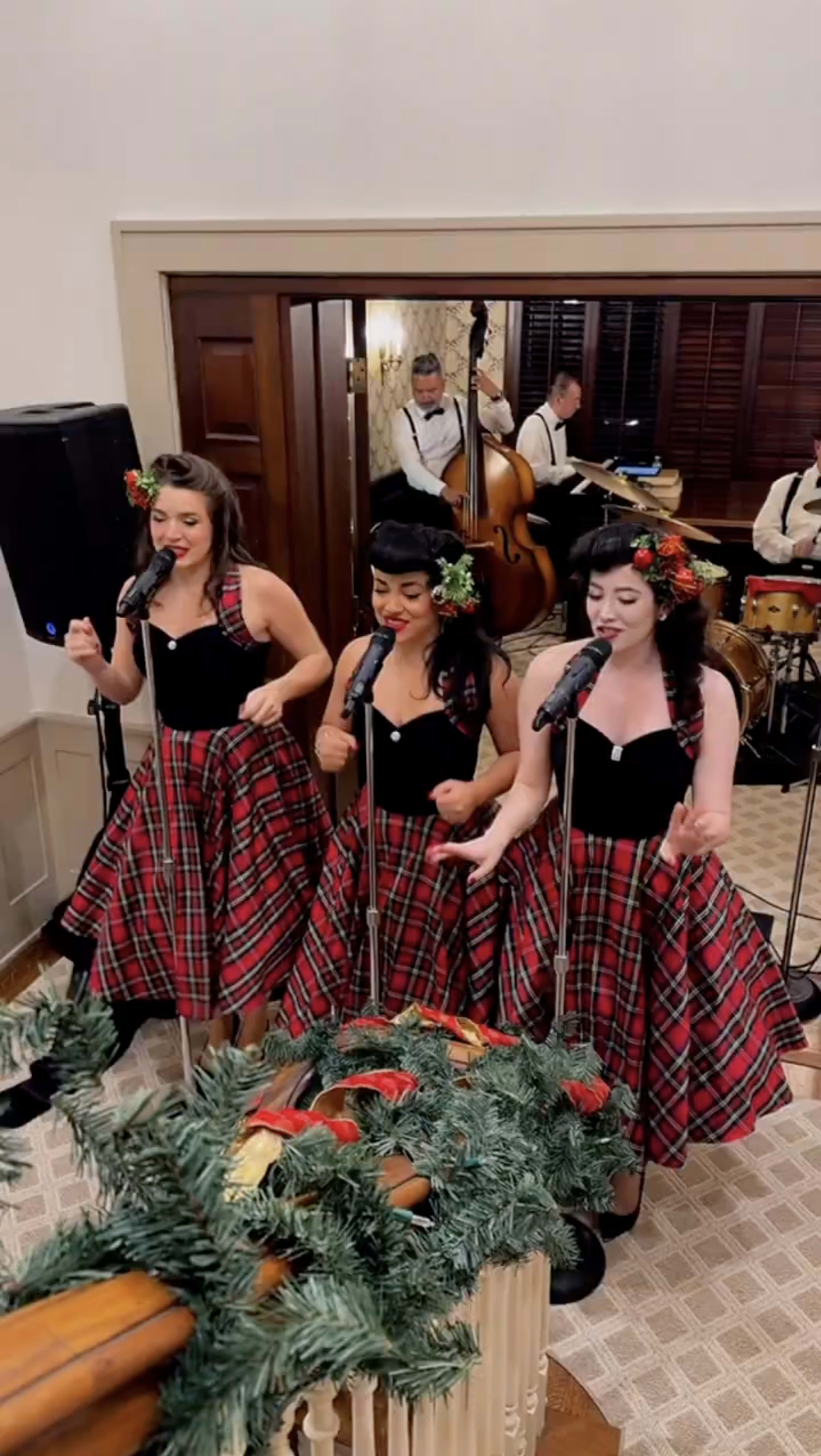 Three women in black tops and tartan skirts perform into microphones, surrounded by festive decorations and a live band in the background.