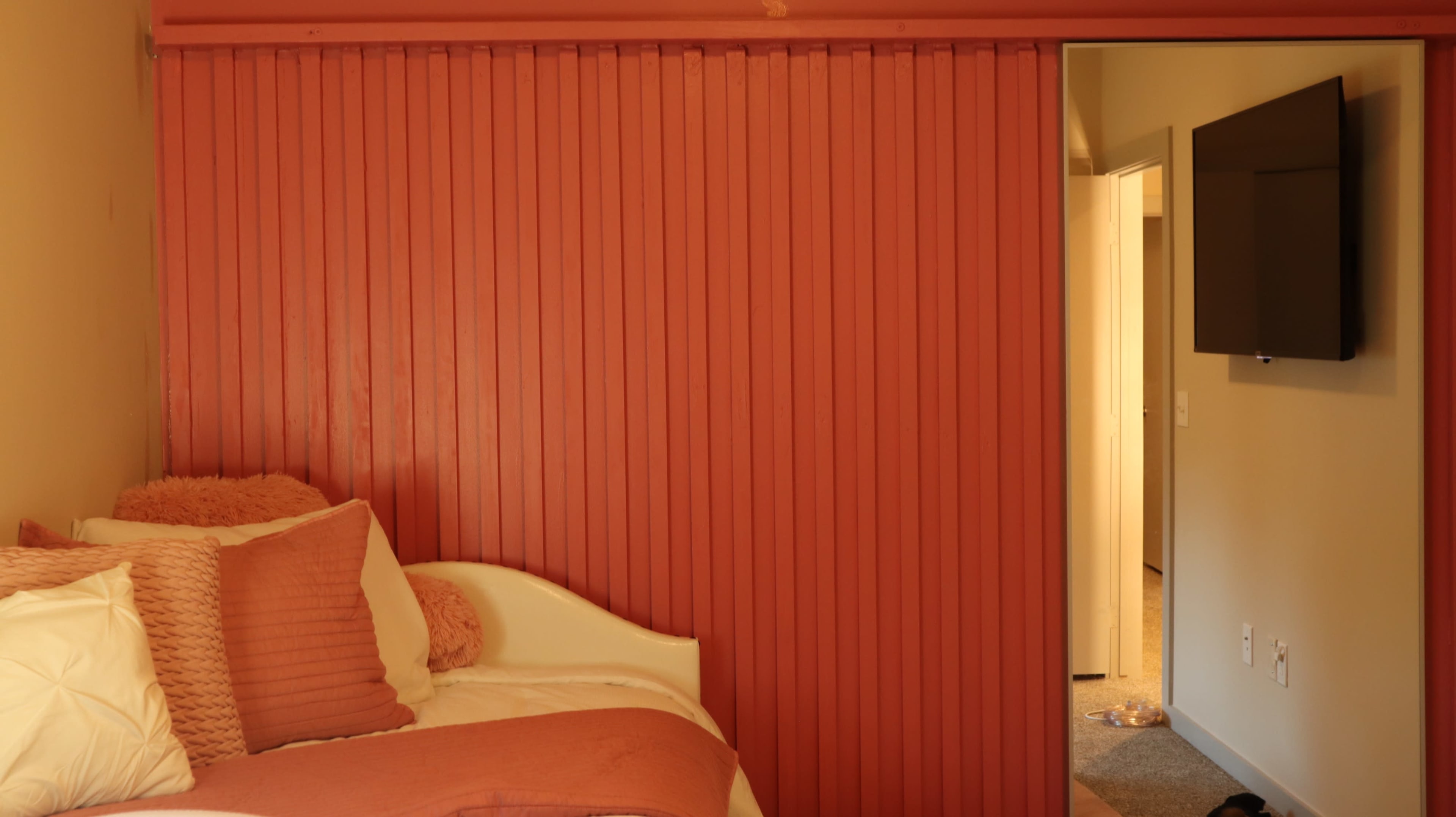 A cozy bedroom features a bed adorned with orange pillows and a wall with vertical wooden paneling, leading to an adjacent room with a light-colored door.