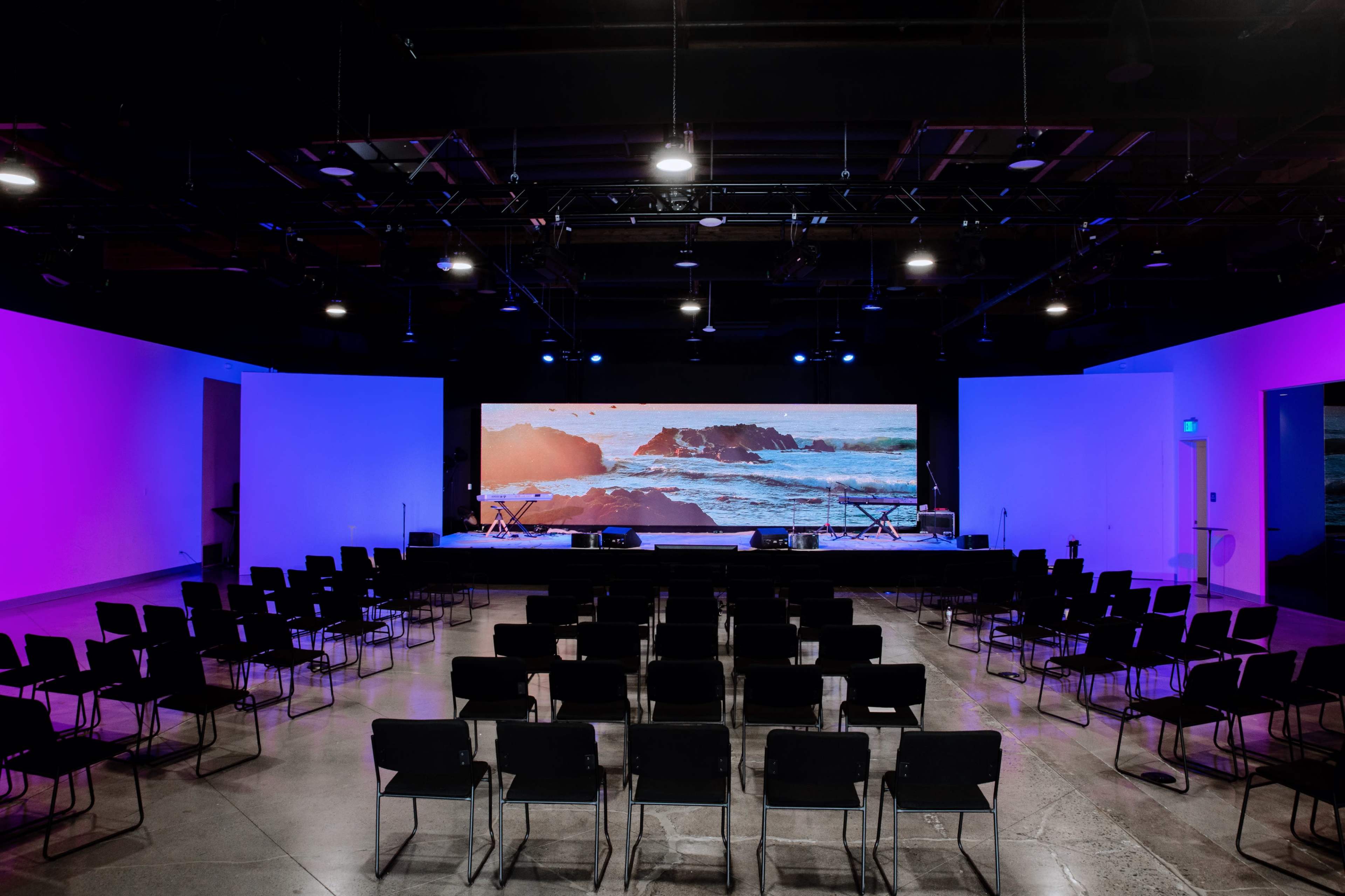An empty auditorium features rows of black chairs facing a large screen displaying a seascape, illuminated by purple lights on the walls.