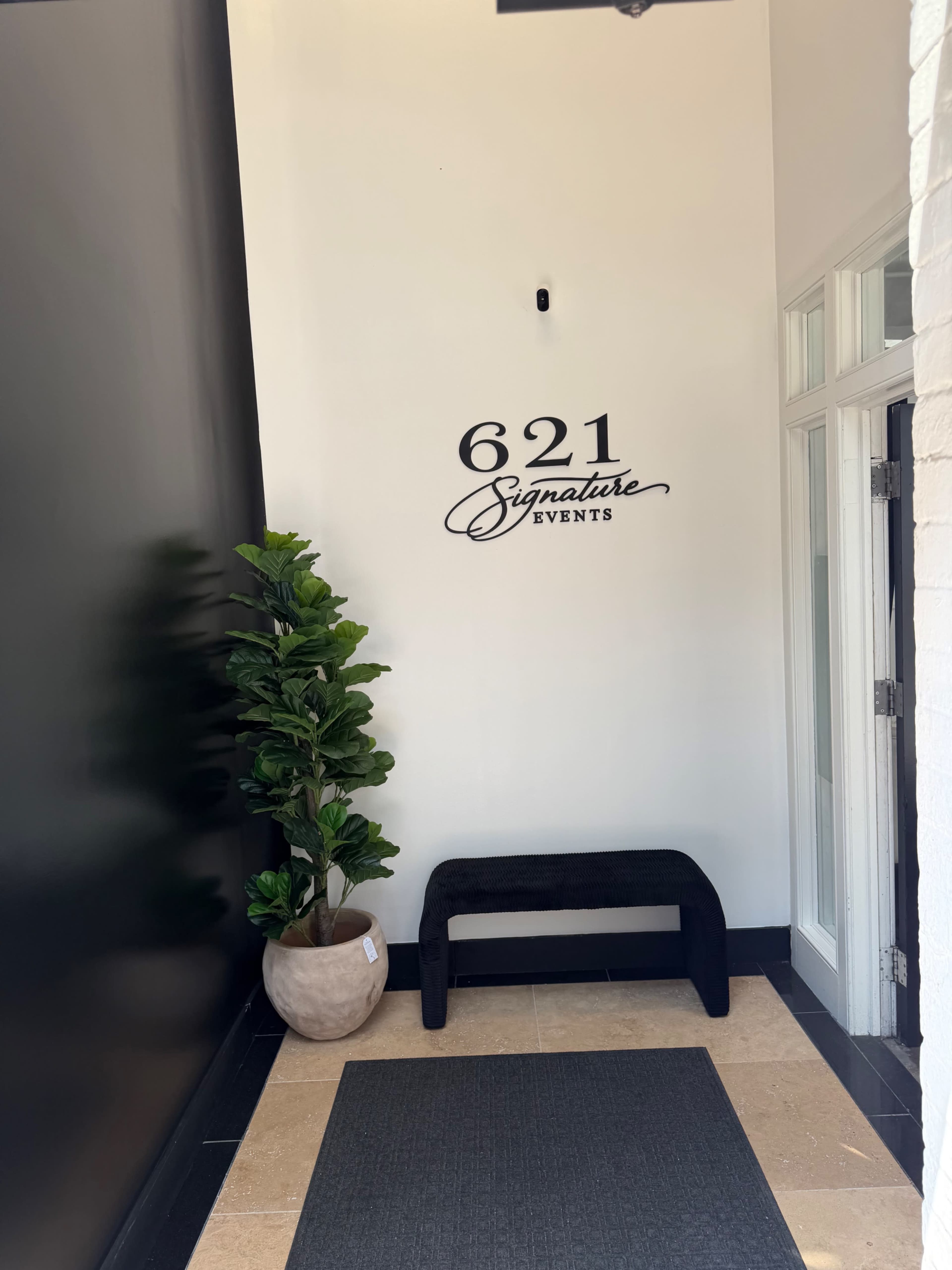The image shows an entrance area with a black wall featuring the text "621 Signature Events," a potted plant, and a black bench on a tiled floor.