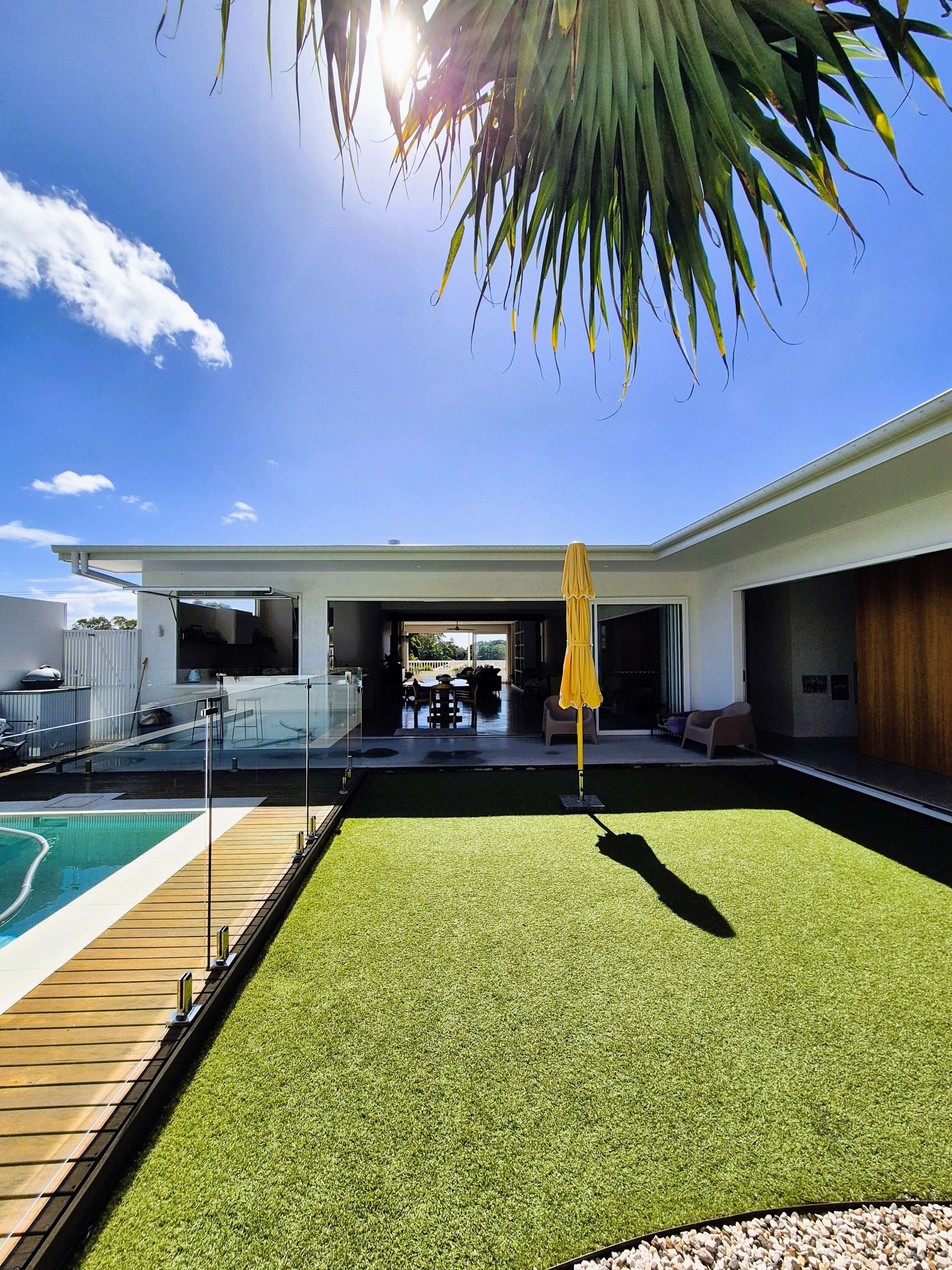 A modern house features a swimming pool, artificial grass, and large windows, with a yellow umbrella standing in the outdoor area.