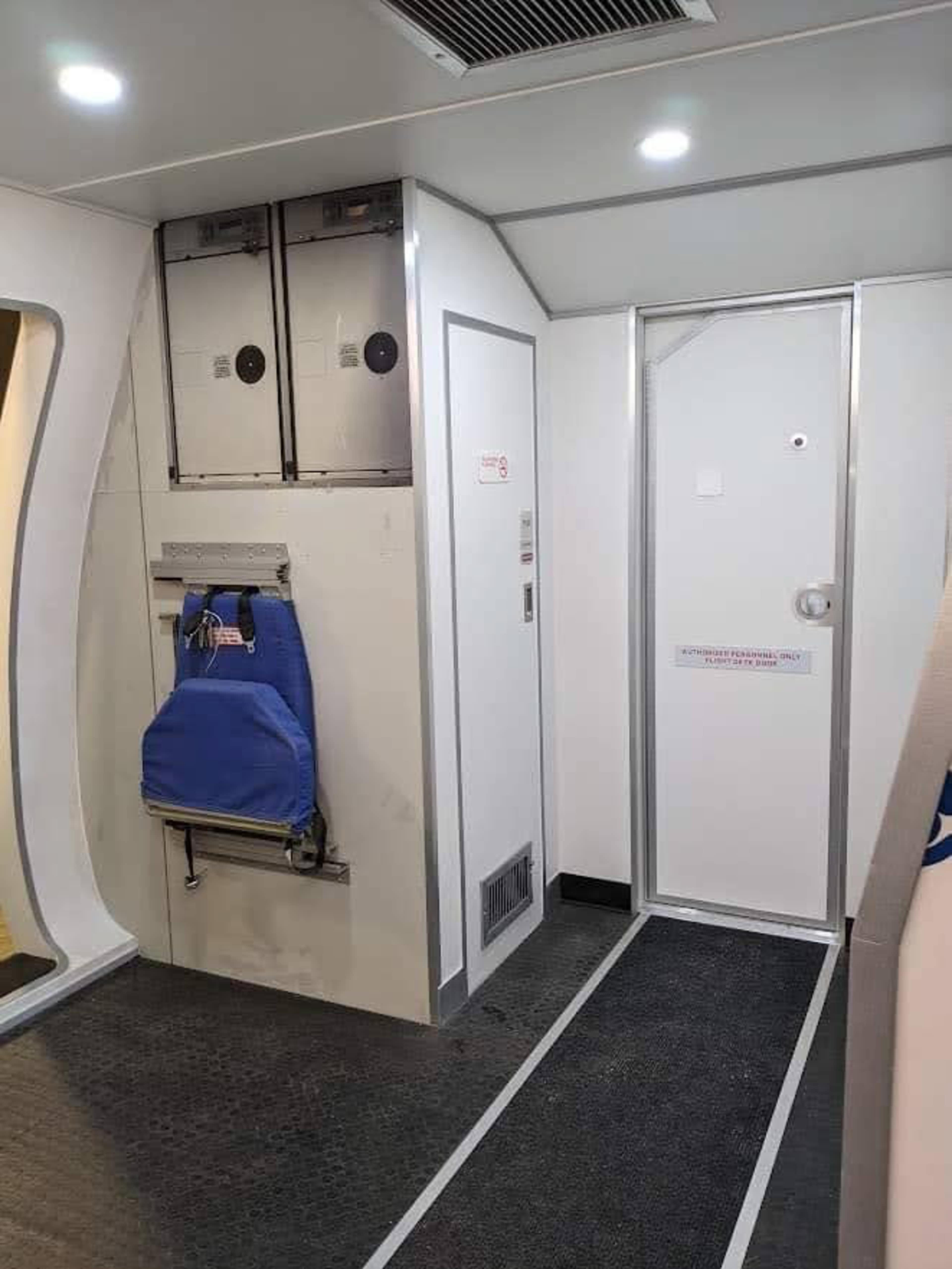 A narrow hallway inside an aircraft, featuring a blue bag hung on the wall and a door leading to a cabin area.