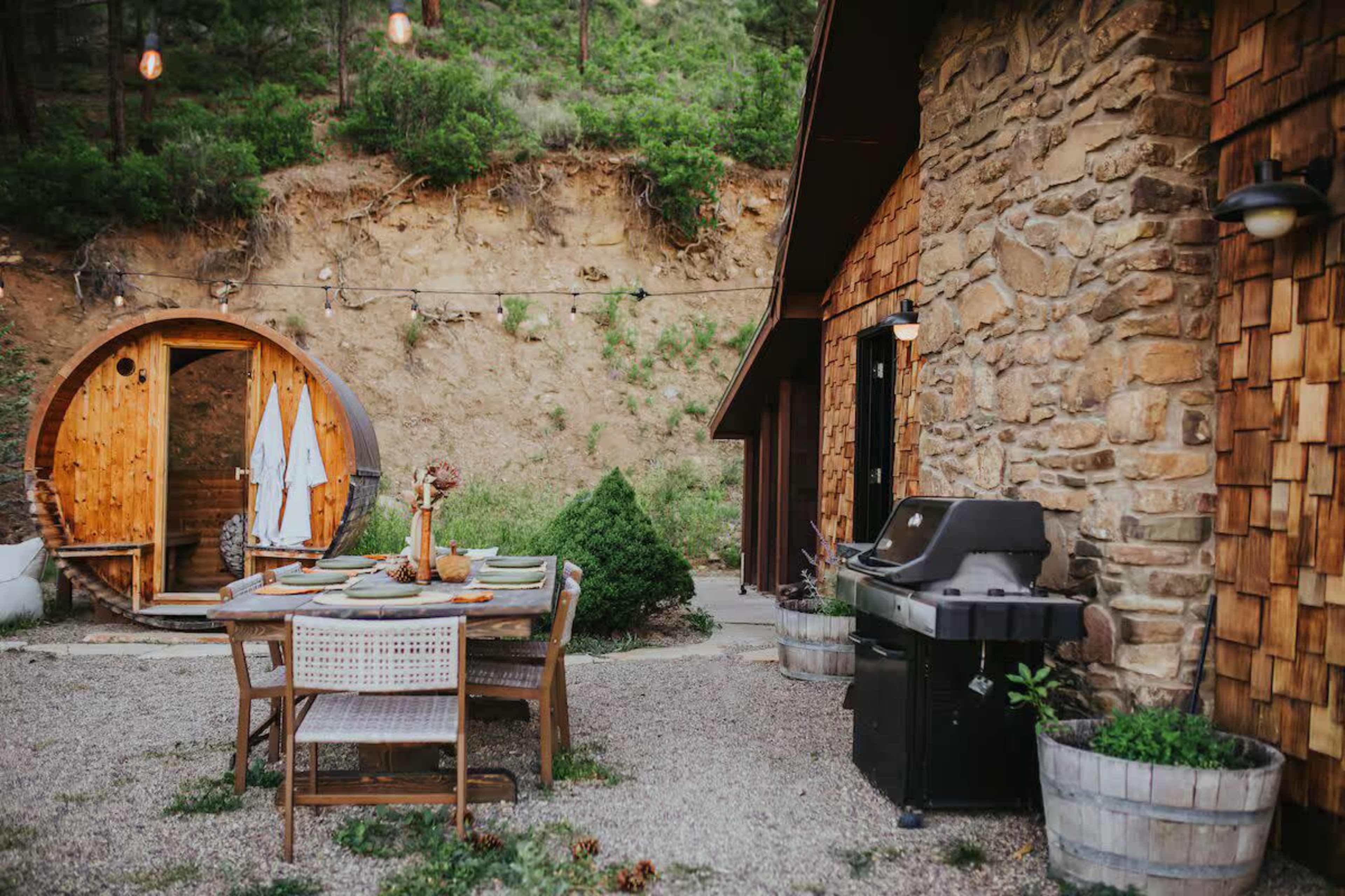 The scene features a rustic outdoor dining area with a wooden table set for a meal, a barbecue grill, and a wooden sauna nearby, surrounded by gravel and greenery.