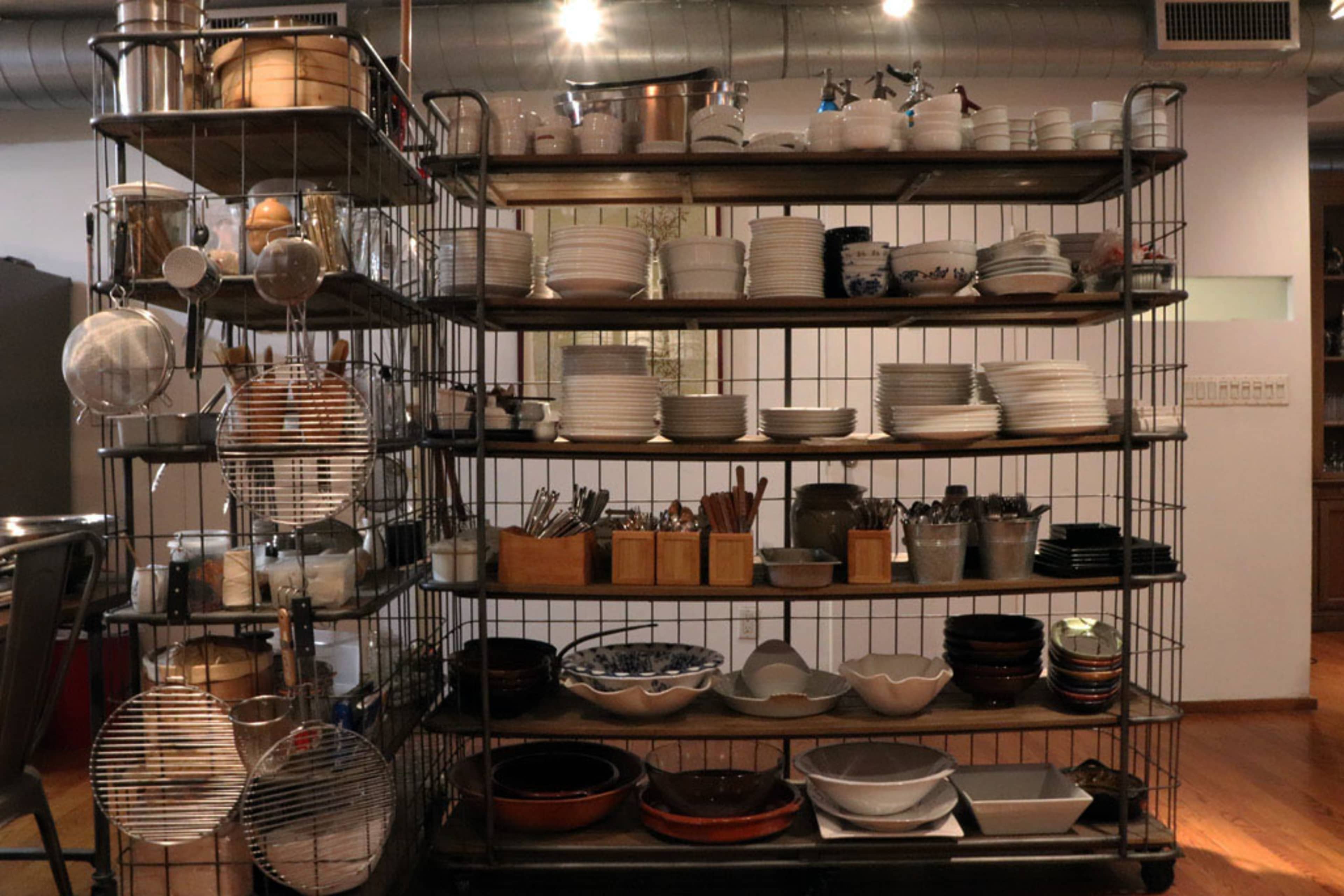 A metal shelving unit displays an assortment of plates, bowls, utensils, and kitchen tools in a kitchen setting.