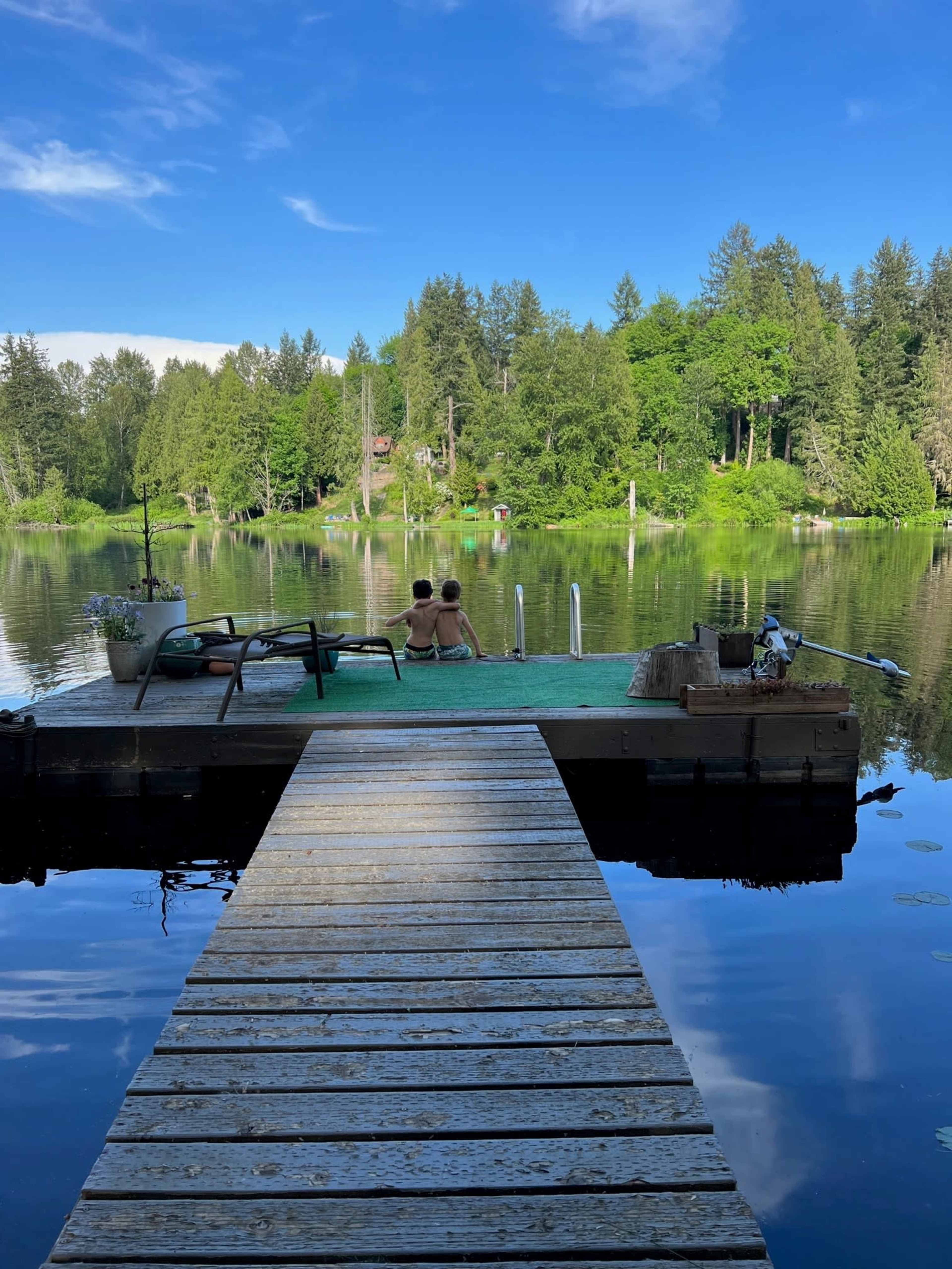 Two people sit on a dock overlooking a calm lake surrounded by trees.