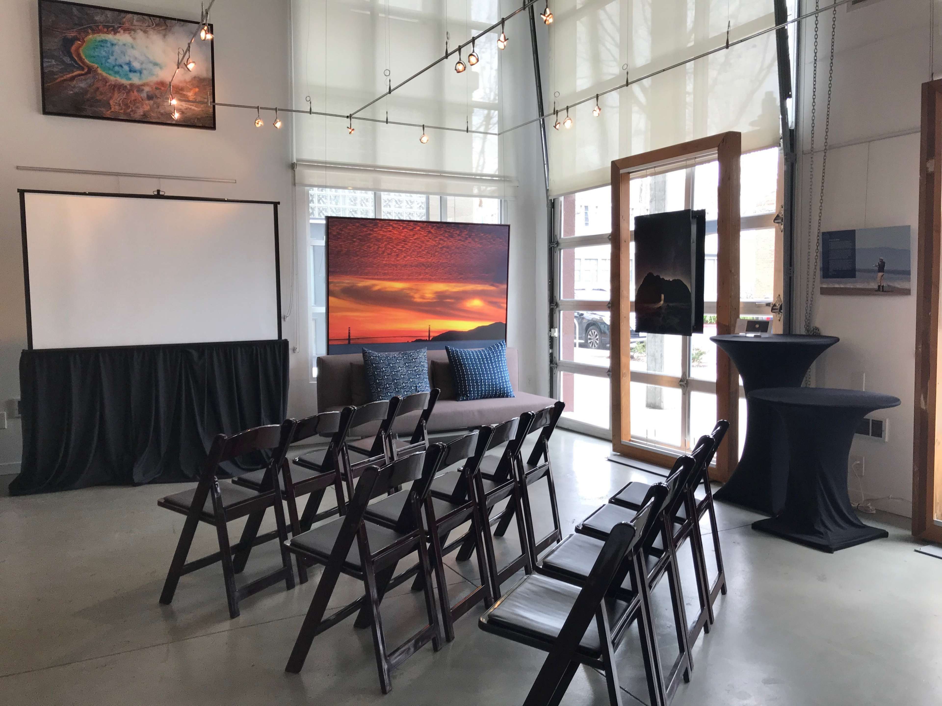 A modern event space set up with rows of folding chairs facing a blank screen, framed by large windows and decorated with artwork on the walls.
