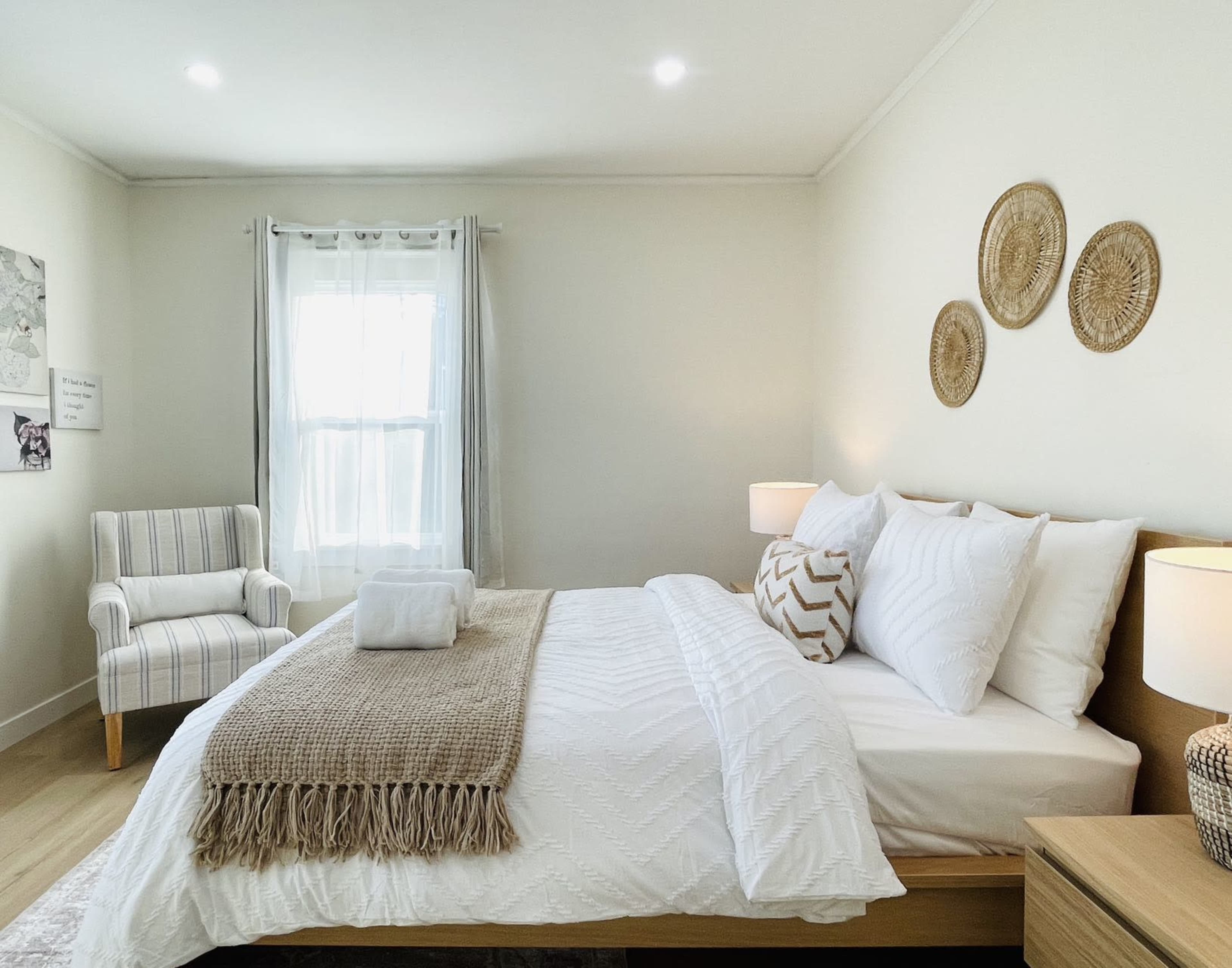 A neatly arranged bedroom features a bed with white linens, decorative pillows, a throw blanket, a chair in the corner, and wall decorations made of woven materials.