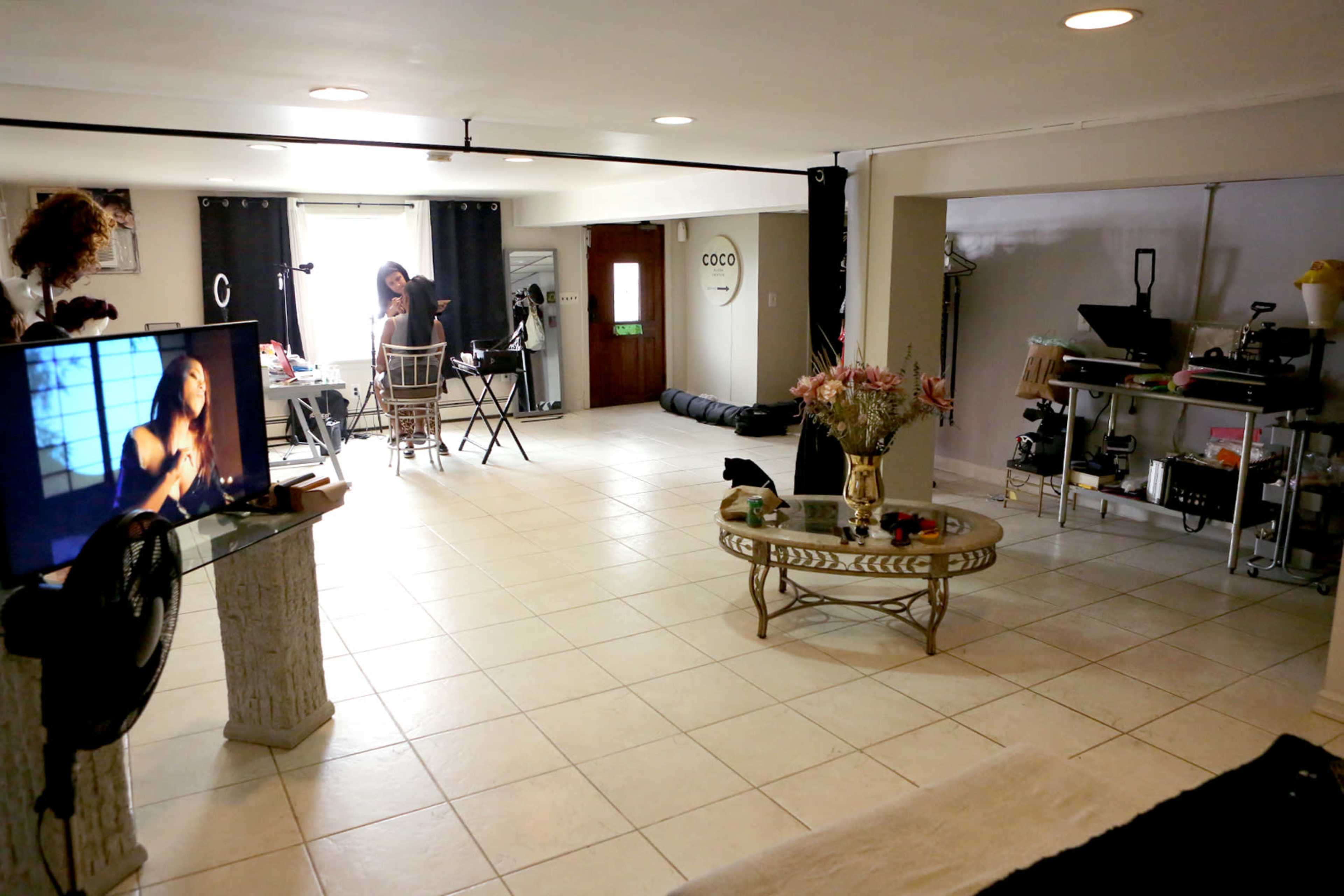 The image shows a spacious room with a tiled floor, featuring a lounging area with a television, a dining setup, and a workspace in the background.