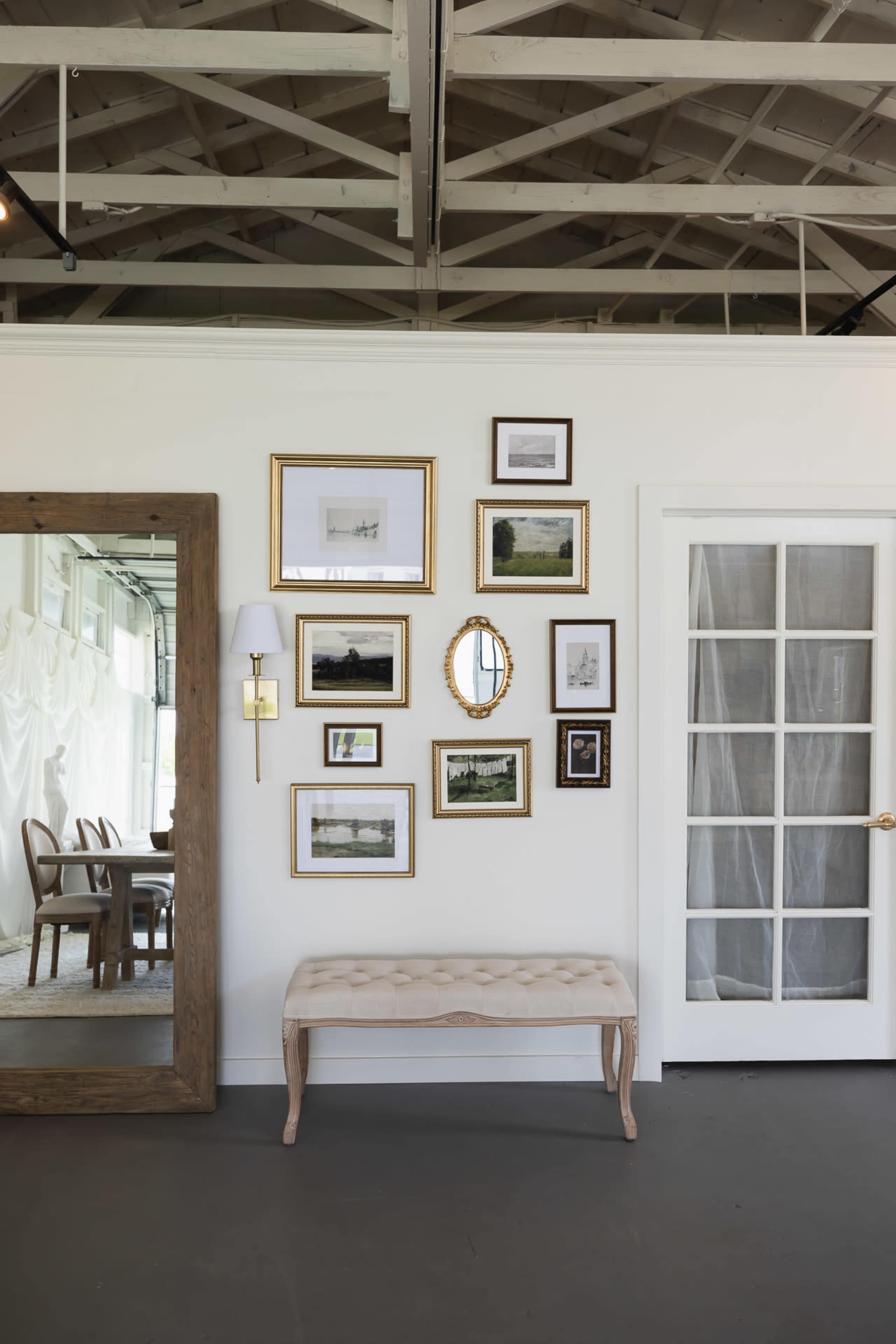 The image shows a well-decorated wall featuring a collection of framed pictures and a round mirror, along with a bench and a door in a bright, spacious room.