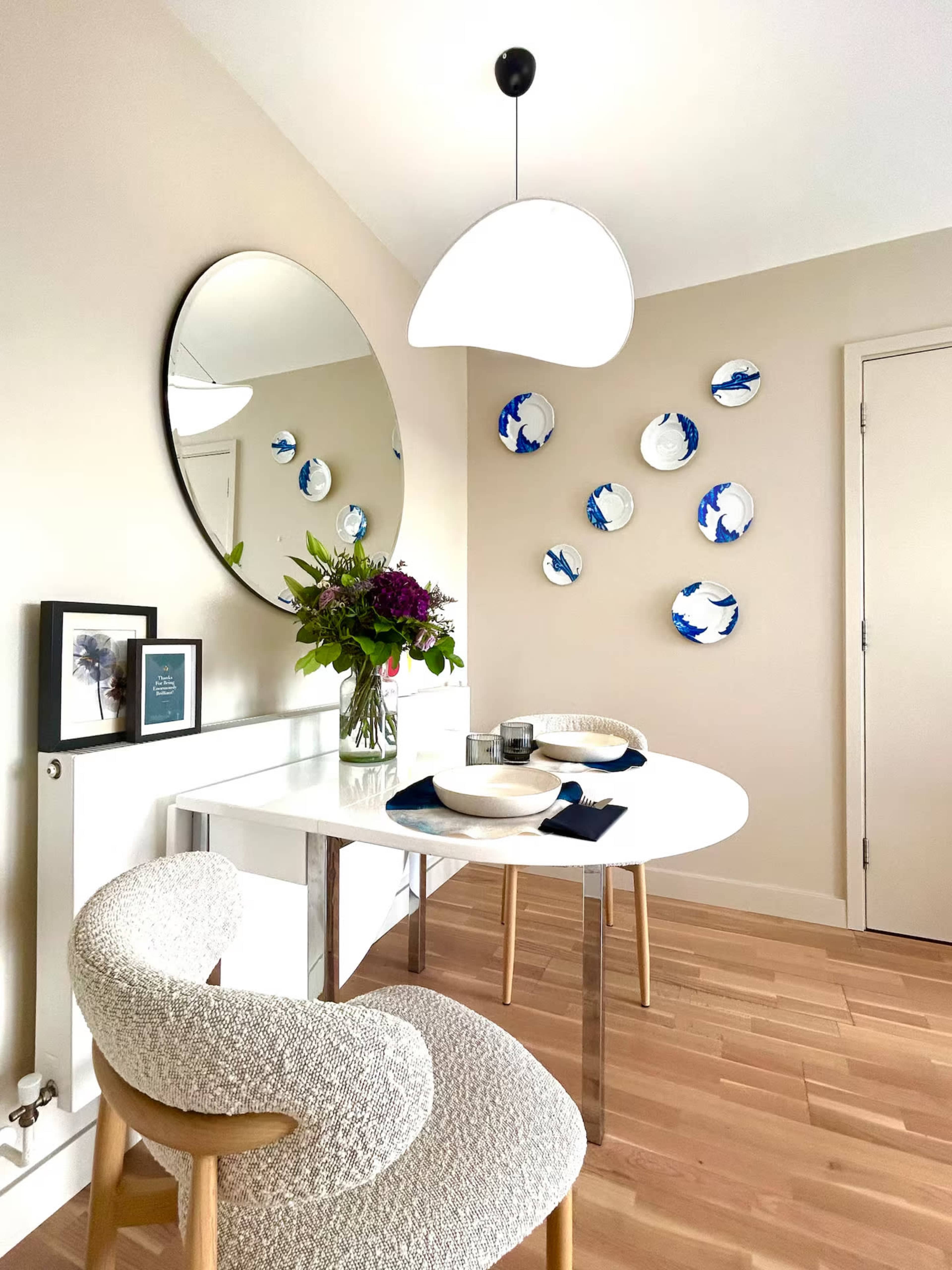The image shows a modern dining area featuring a round table with two chairs, a large circular mirror on the wall, and decorative plates displayed on the adjacent wall.