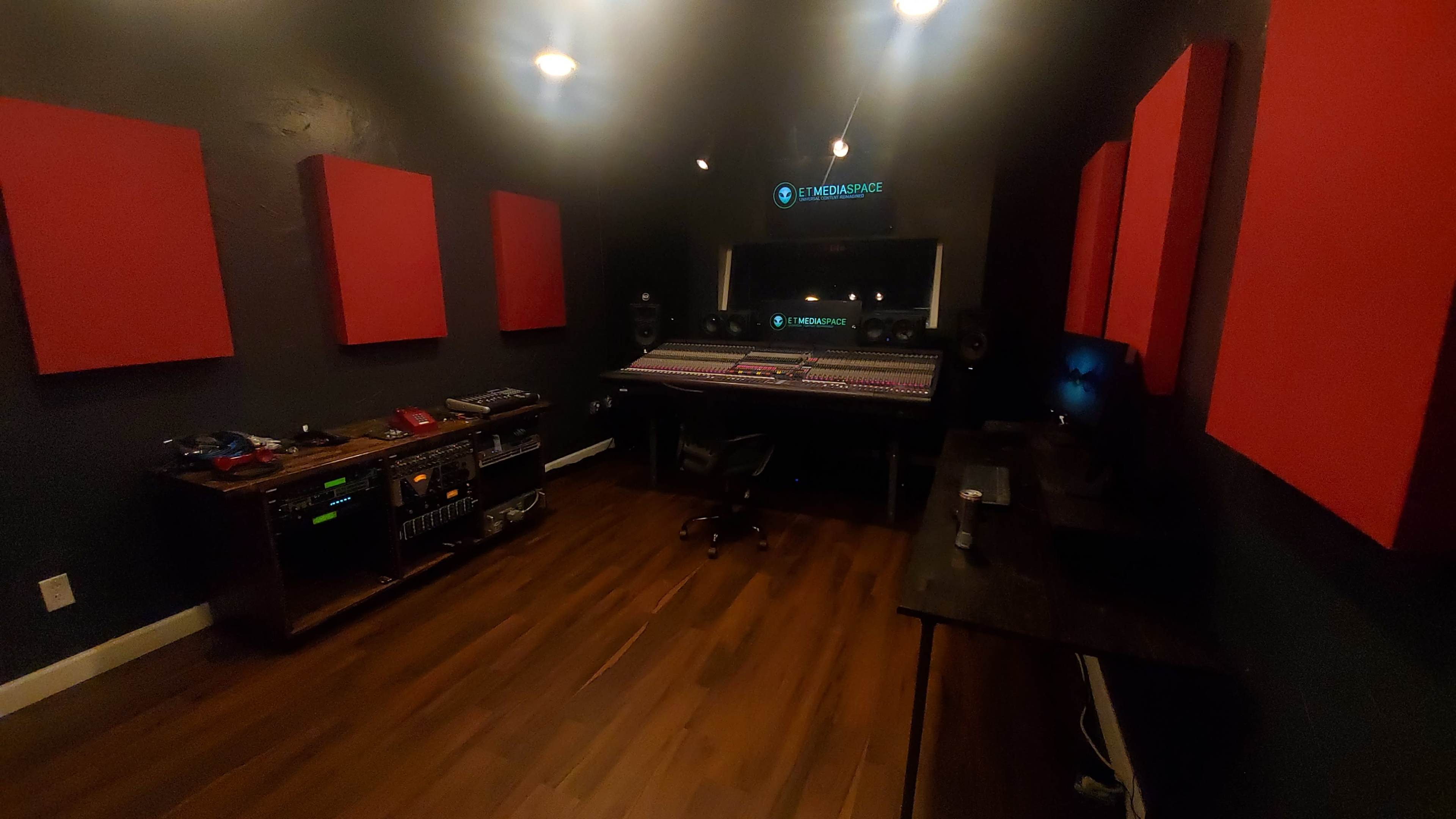 The image shows a music production studio with a large mixing desk, speakers, and red acoustic panels on the walls, featuring wooden flooring and various audio equipment.
