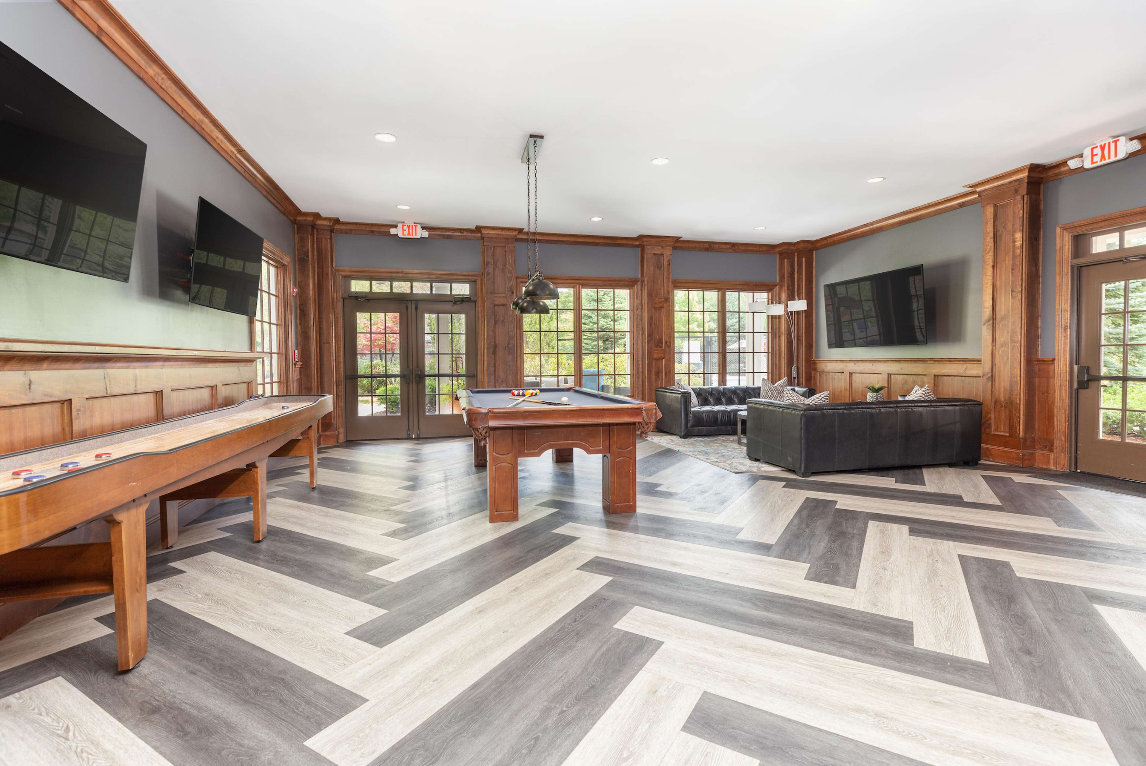 A spacious lounge area featuring a billiard table, a shuffleboard table, and a seating arrangement with a black sofa, all set against a backdrop of large windows and wooden paneling.